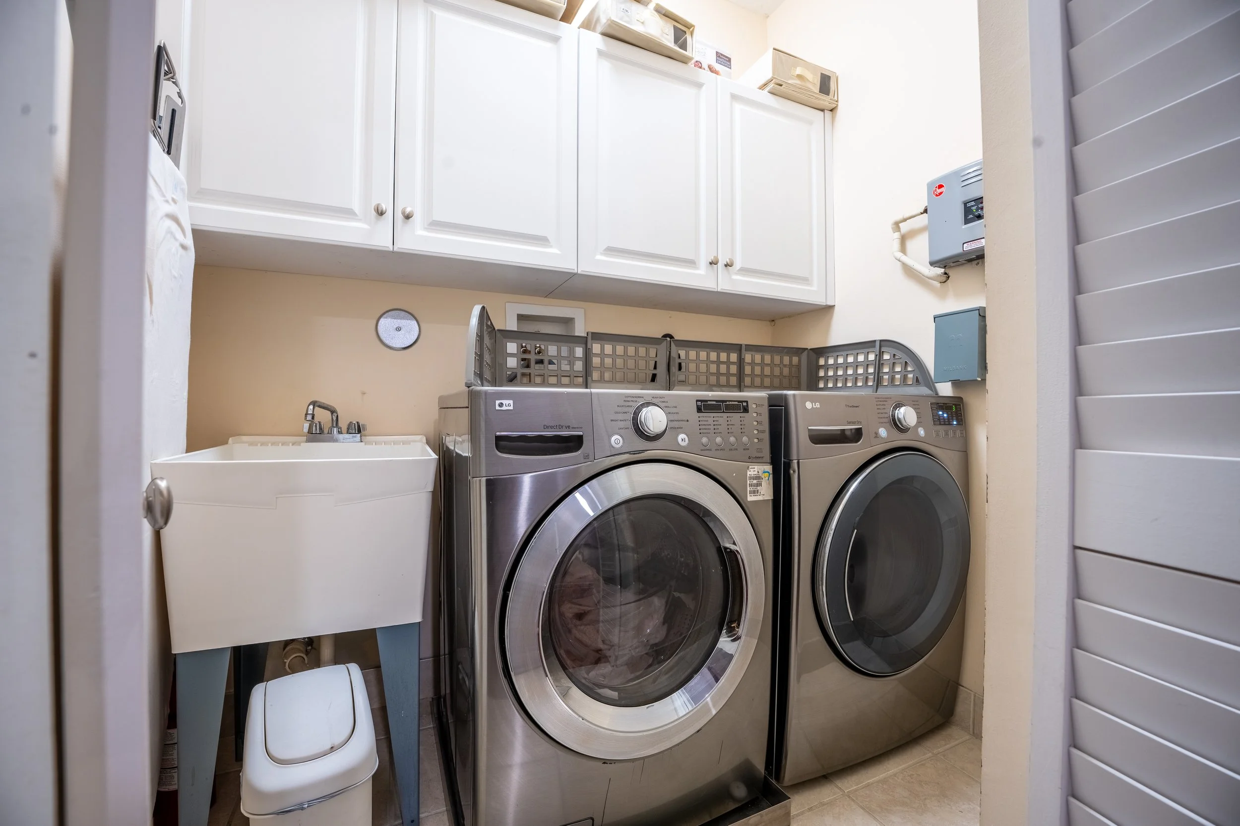 A laundry room with white cabinets above a washing machine and dryer. There is a small white laundry basket or container on the left, and a blue stand underneath the sink with a faucet. On top of the cabinets, there are tan boxes or packaging, and th