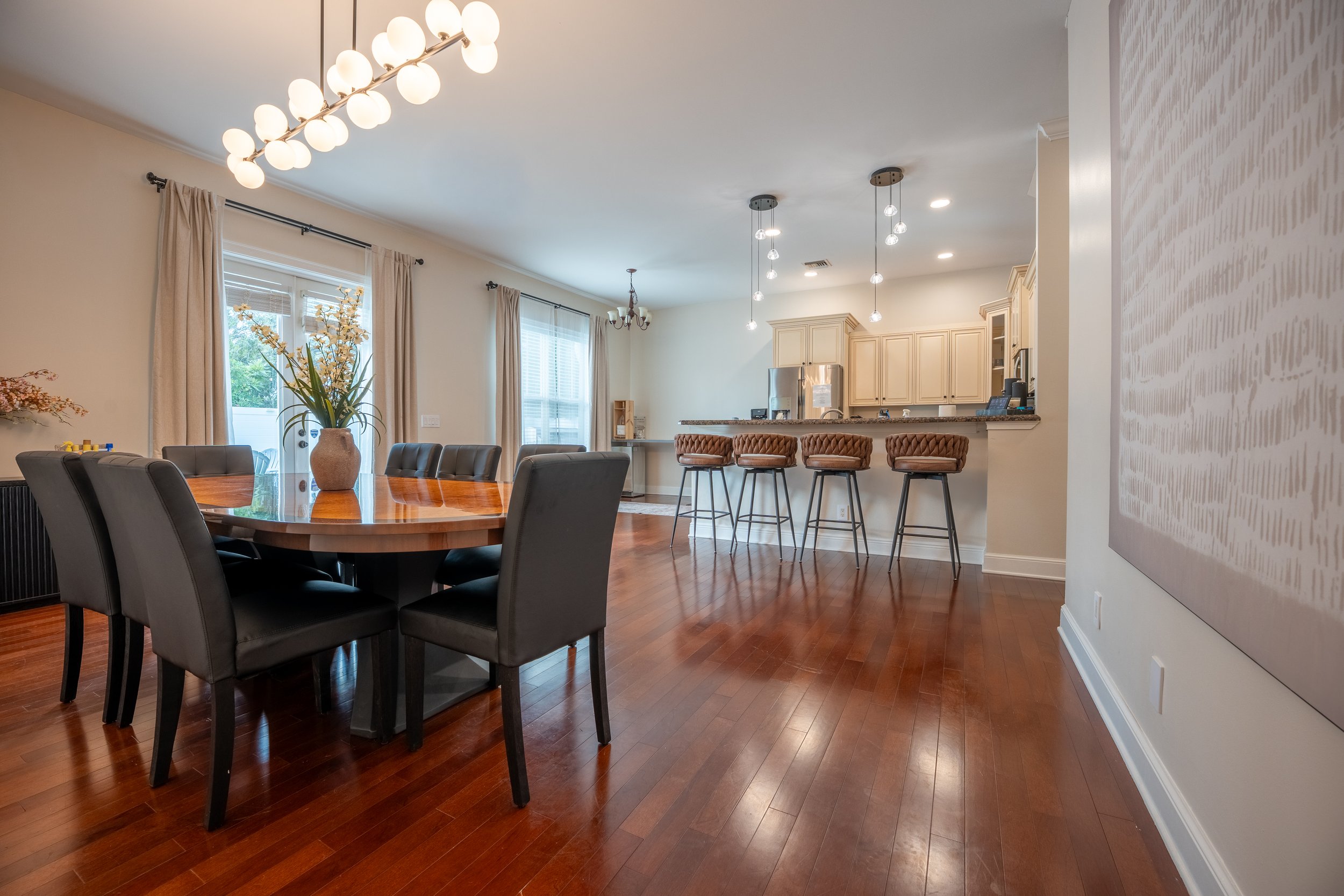 A spacious dining area with a wooden table and six black chairs. The room has large windows with beige curtains, a modern chandelier, and wooden flooring. The kitchen in the background features cream-colored cabinets, a stainless steel refrigerator, 