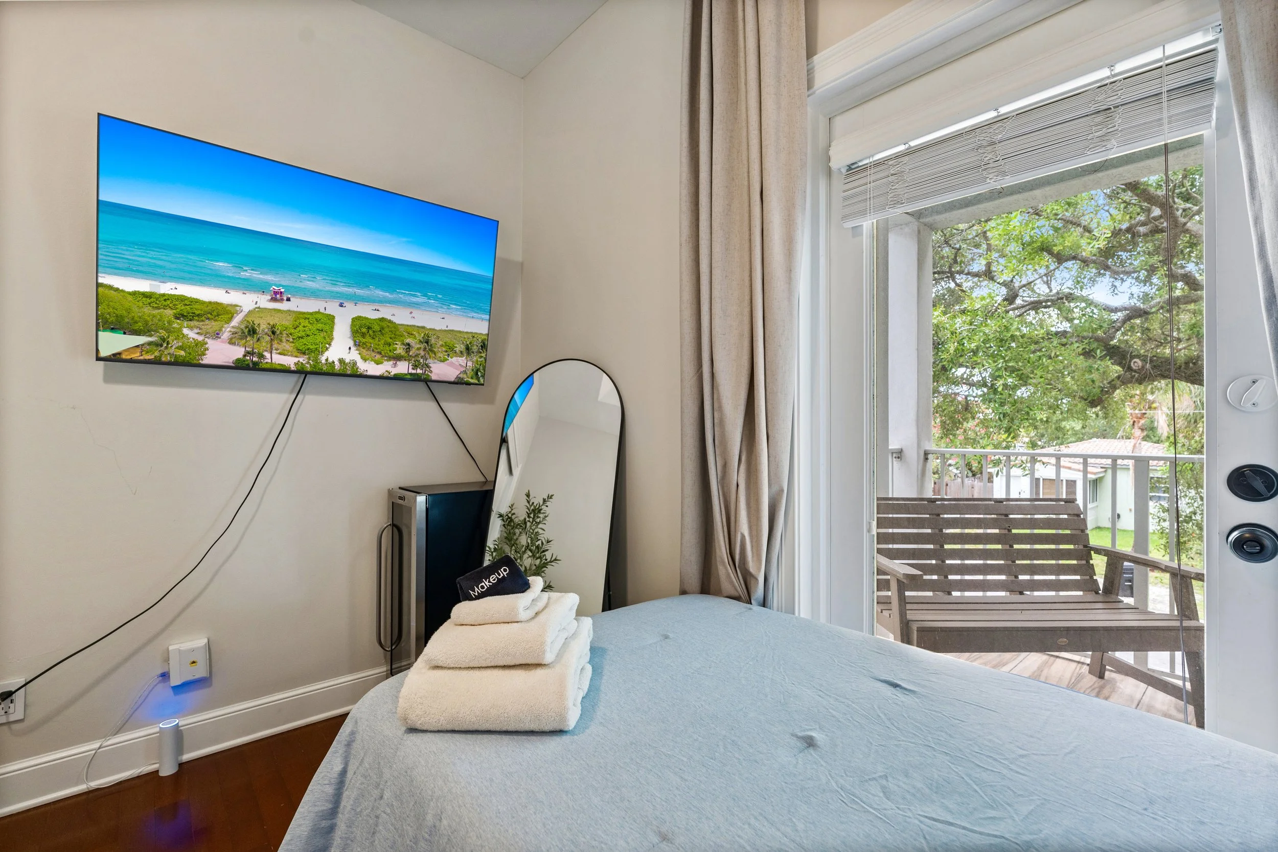 A bedroom with a wall-mounted TV showing a beach scene, a bed with folded towels, a small mirror, and a sliding door leading to a balcony with a wooden chair. Outside, green trees and a blue sky are visible.