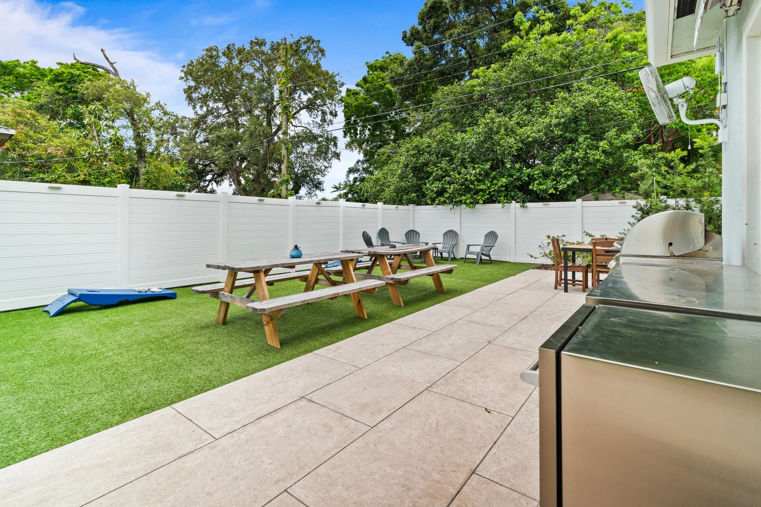 Backyard patio with green artificial grass, white fence, picnic table, plastic chairs, a grill, and trees in the background.