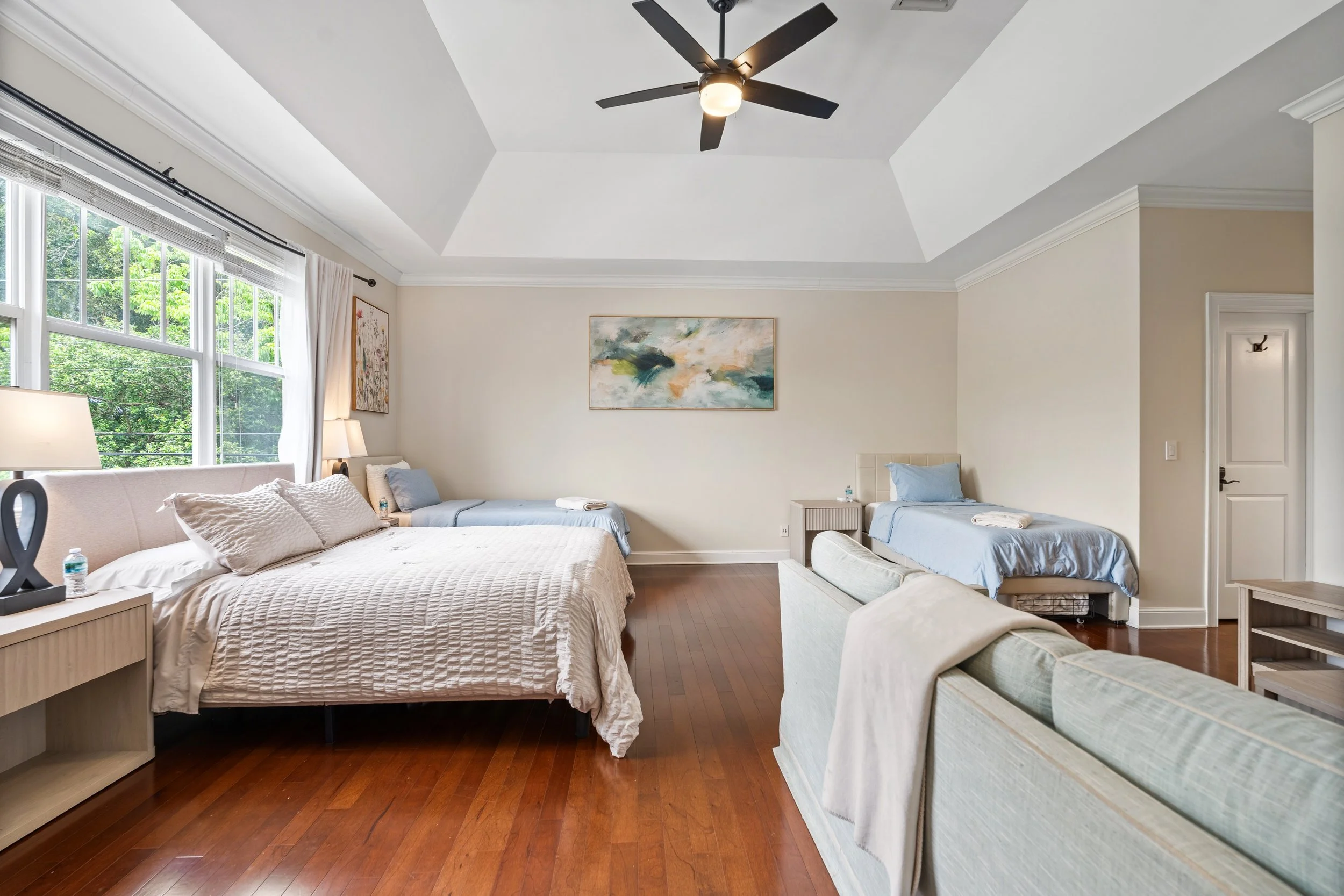 A spacious bedroom with a large bed, a beige and white striped sofa, a ceiling fan, and artwork on the walls. Large windows let in natural light and show greenery outside.