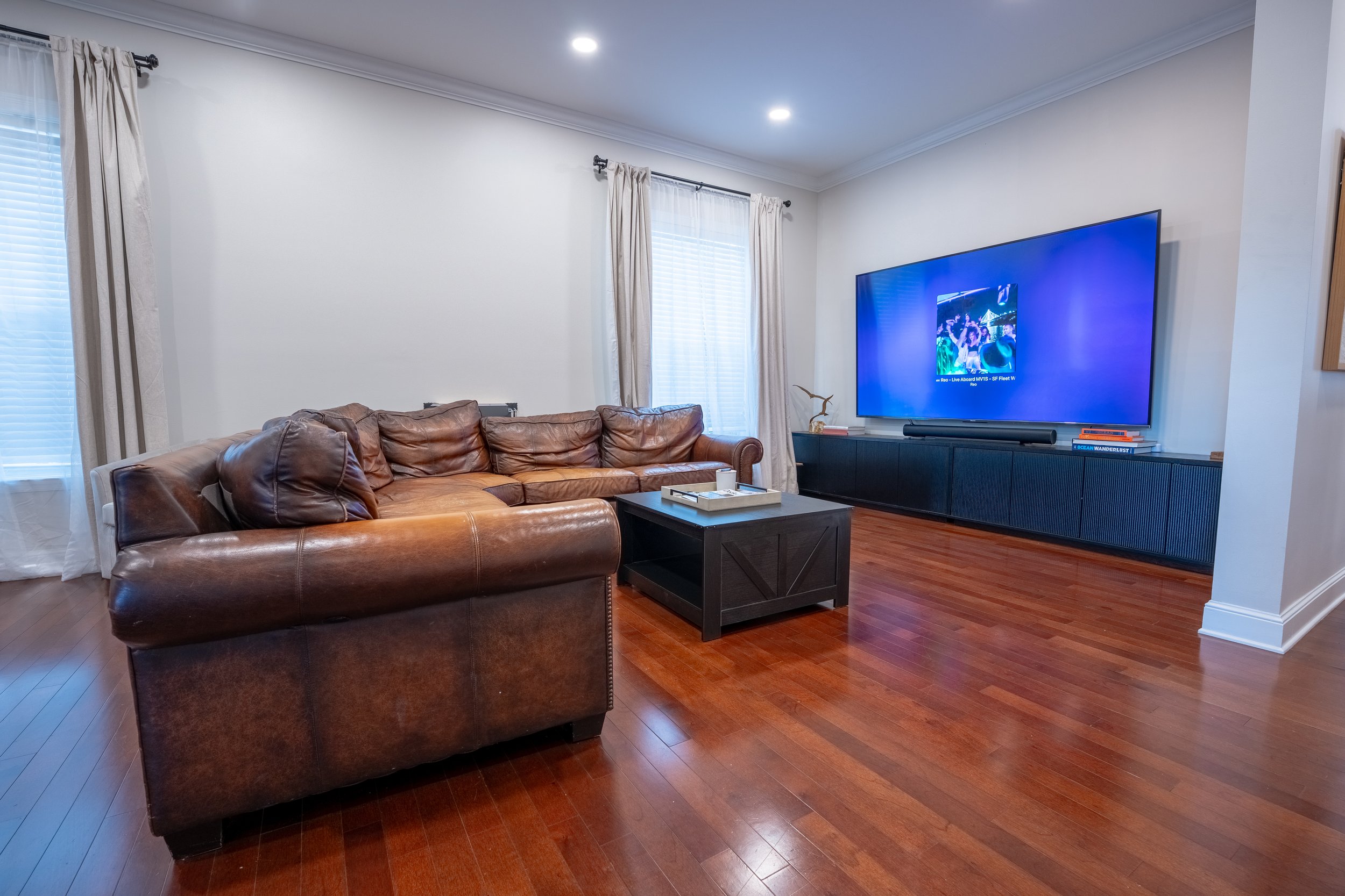 Living room with a leather sofa, window with beige curtains, a large flat-screen TV on a black entertainment unit, and hardwood flooring.