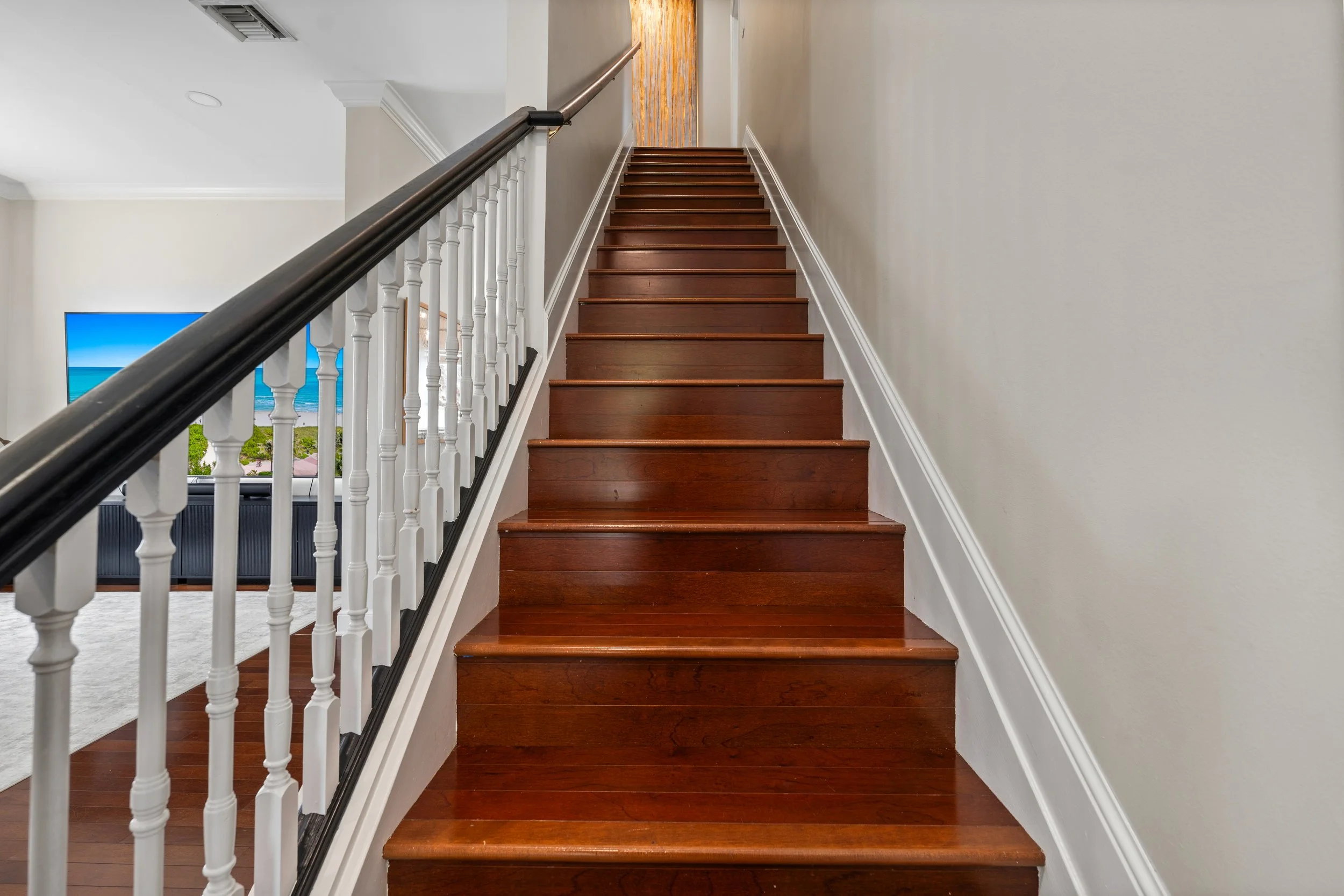 Indoor staircase with wooden steps, white walls, and a black handrail on the left side, leading up to a landing with an orange-colored rug.