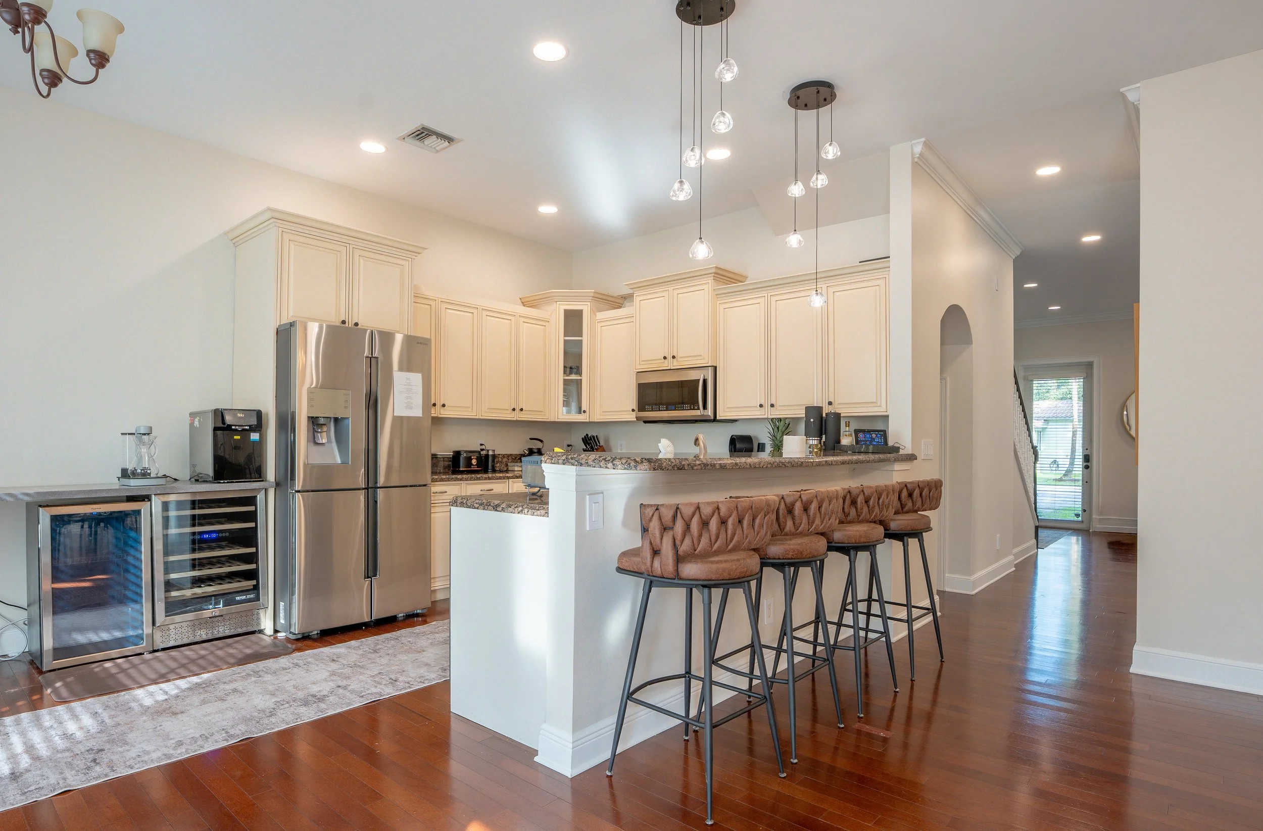 Kitchen area with white cabinets, stainless steel refrigerator, microwave, and a granite countertop island with four brown tufted barstools. Recessed lighting and a modern chandelier with hanging glass bulbs are above the island. The floor is hardwoo