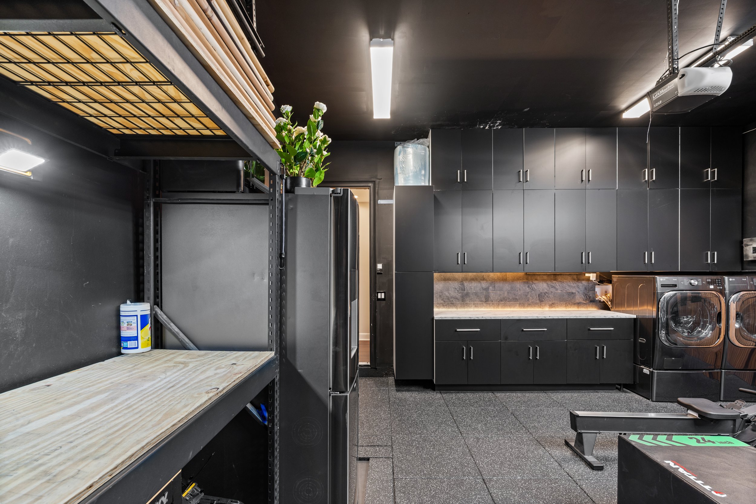 A room with black walls and cabinets, a water dispenser, a potted plant, and empty shelves with a roll of paper towels. The floor is black and the ceiling has fluorescent lights.