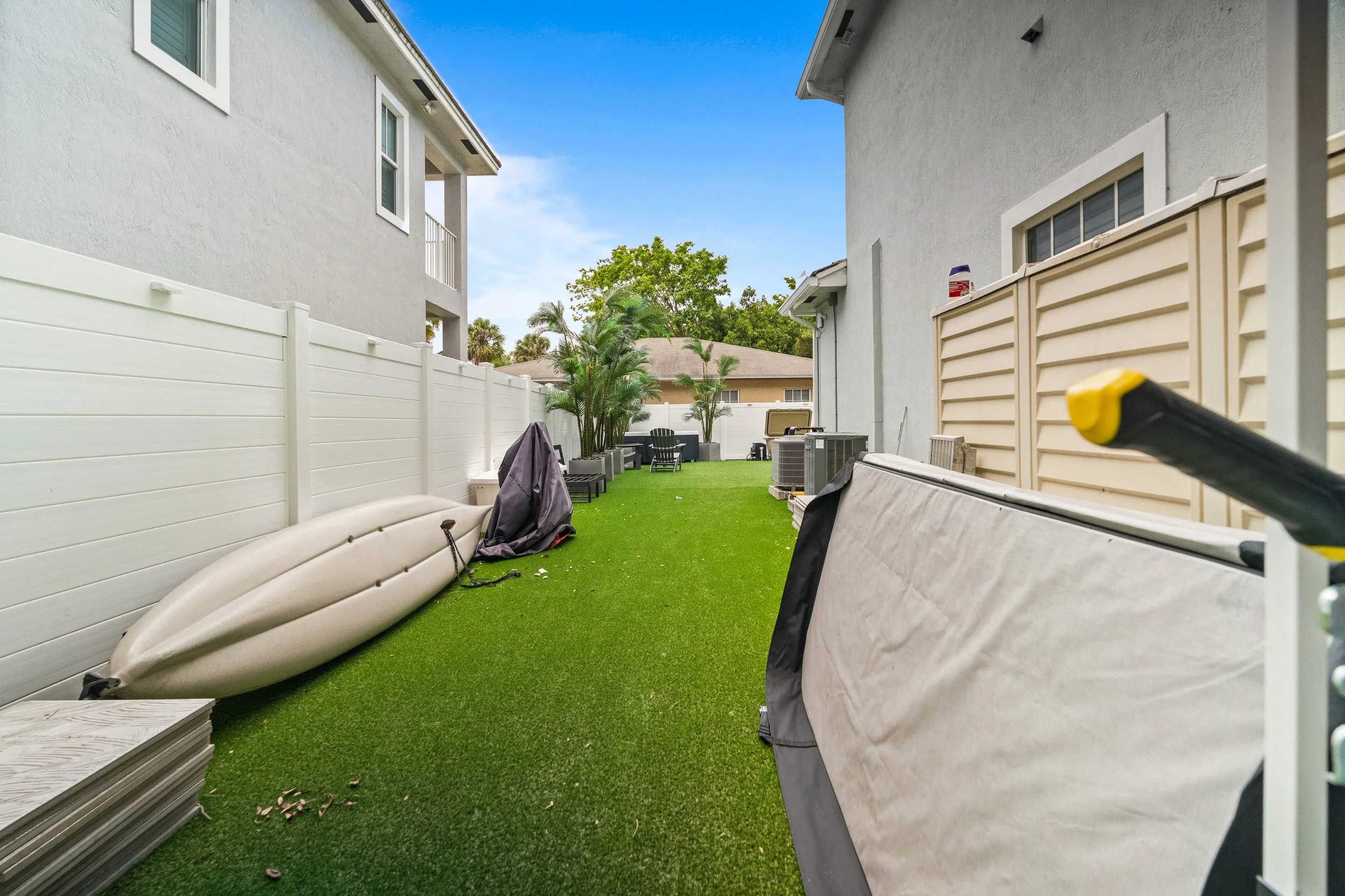 A backyard with artificial grass, white fences, and various outdoor furniture including paddleboards, a grill, and lounge chairs. There are tall trees in the background, and the sky is blue with some clouds.