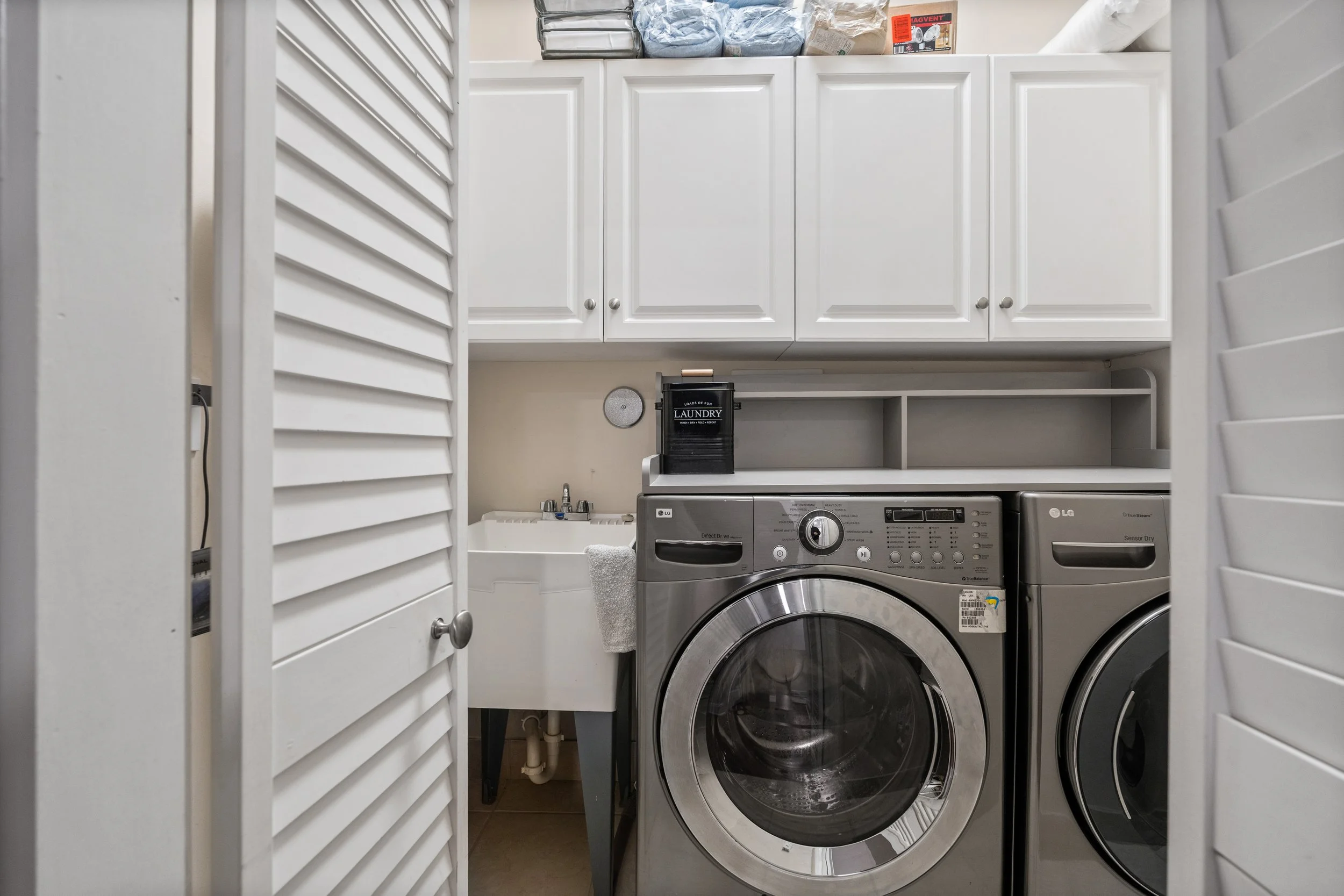 Laundry room with white cabinets, a front-loading washing machine, and a dryer, with shelves on top holding laundry supplies. Louvered doors partially open, revealing the laundry area.