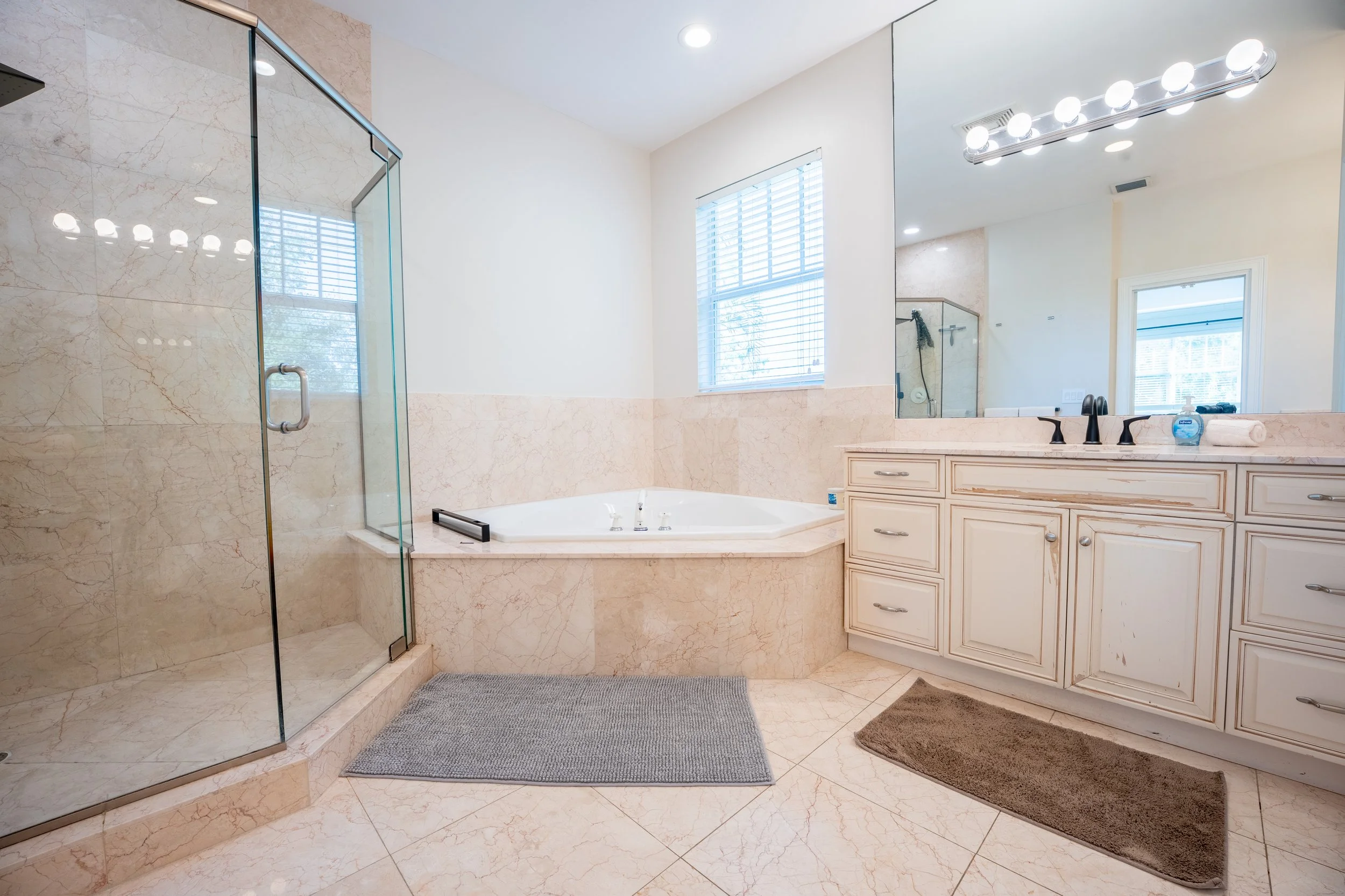 A spacious bathroom featuring a glass-enclosed shower, a bathtub with a marble surround, a large mirror above a cream-colored vanity with multiple drawers, a modern black faucet, a window with white blinds, and various towels and toiletries on the co