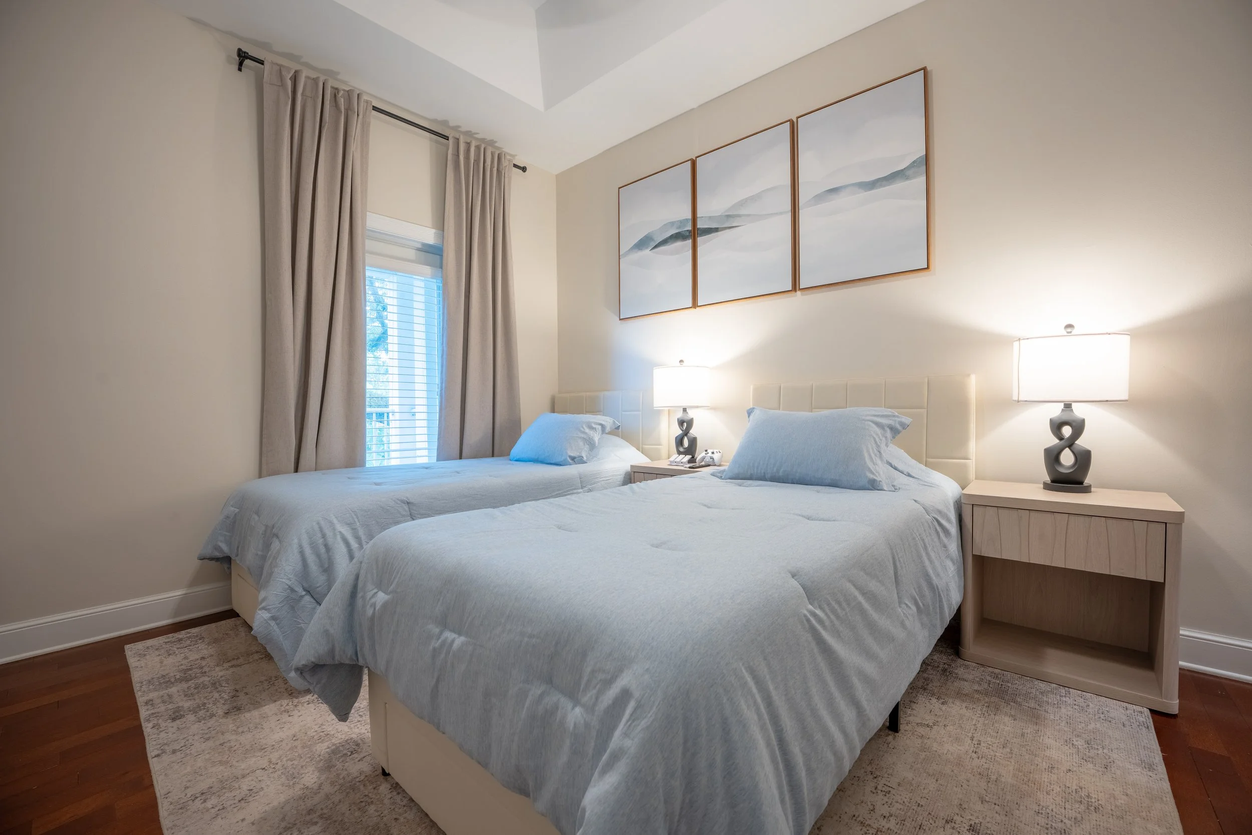 Two twin beds with light blue bedding and pillows in a bedroom. There are nightstands with lamps on either side of the beds. A window with beige curtains and blinds is on the left side, and a triptych painting of mountains hangs above the headboards.