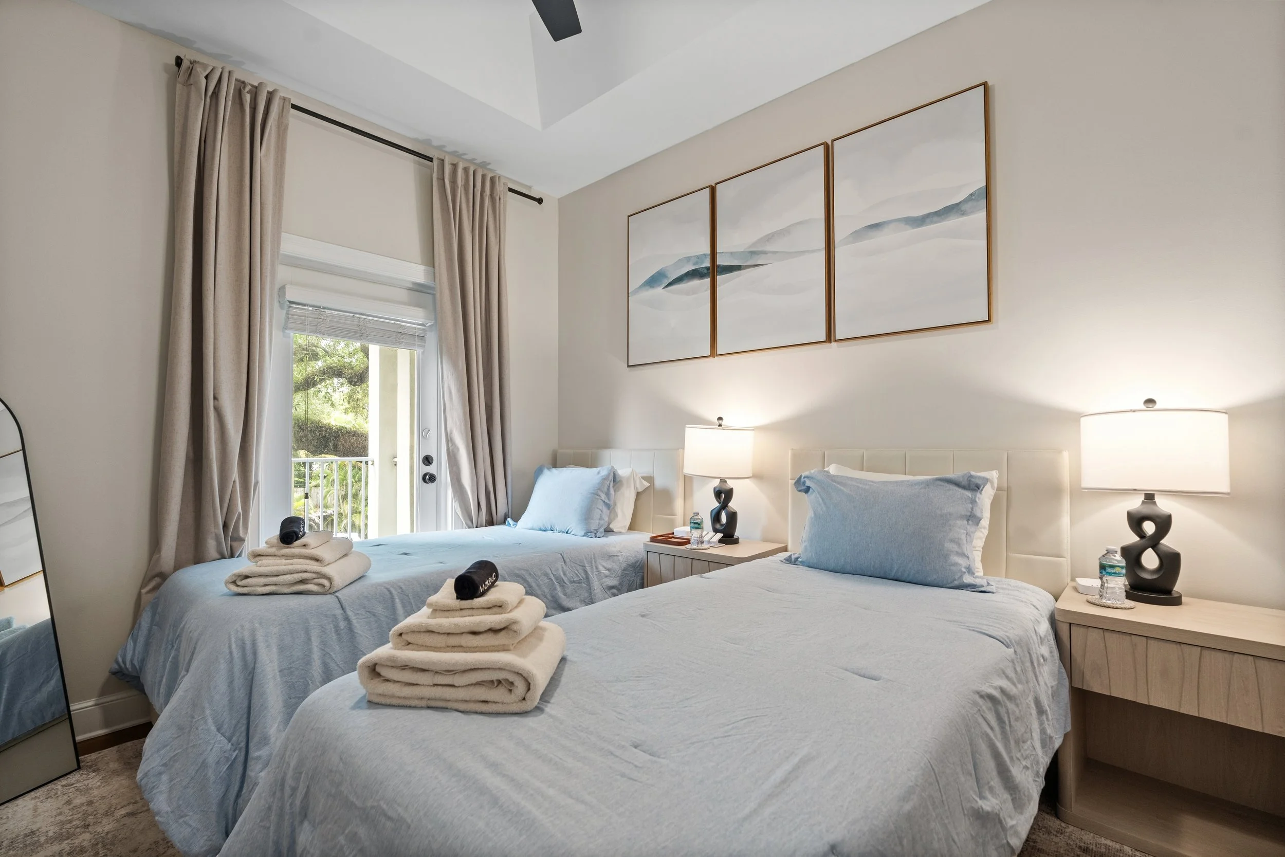 A hotel room with two beds, each with light blue bedding and pillows. Towels are neatly folded on the beds. There is a window with beige curtains, and a balcony door that shows greenery outside. Two matching nightstands with lamps are placed beside t
