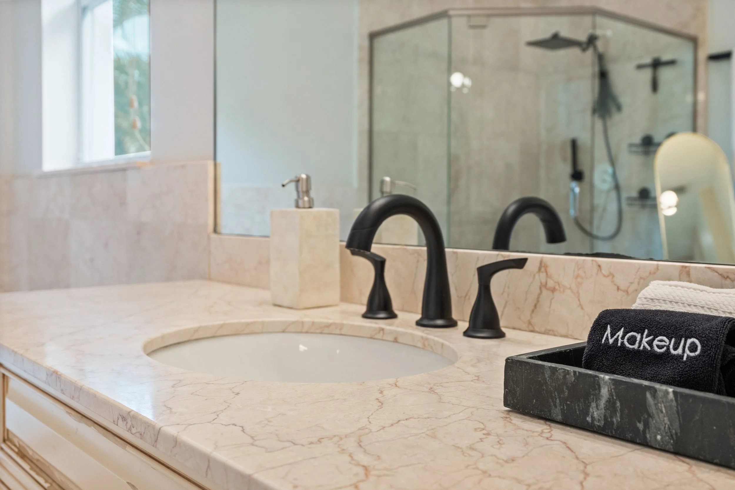 A bathroom countertop with a marble surface, black faucets, a soap dispenser, and a black tray holding a towel with the word 'Makeup' embroidered on it. A mirror is mounted above the countertop, reflecting a glass-enclosed shower with a showerhead an