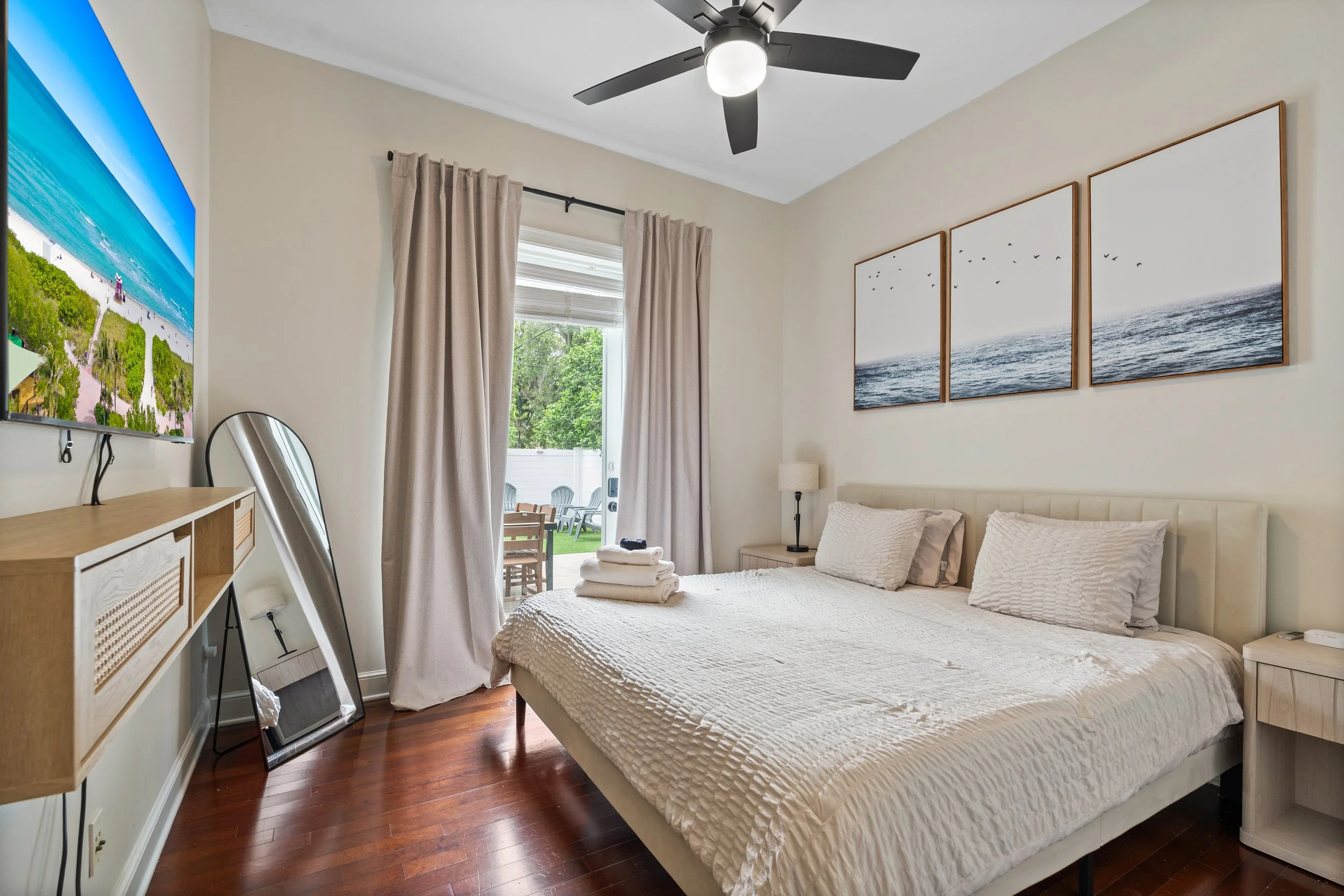A bedroom with a bed, nightstand, window with curtains, wall art, a ceiling fan, a mirror, and a TV.