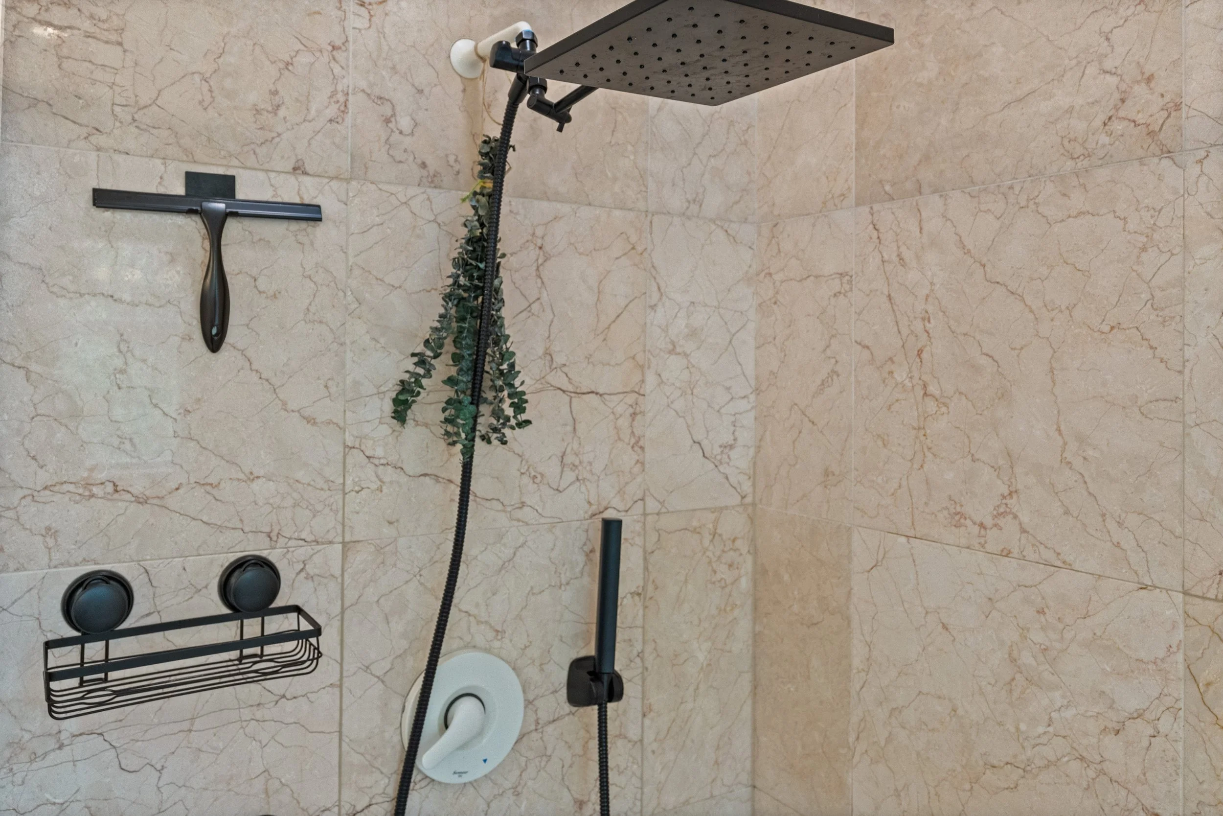 A marble shower wall with a black showerhead, a handheld shower wand, a wall-mounted soap dish, a small shelf with toiletries, and some decorative greenery hanging from the showerhead.