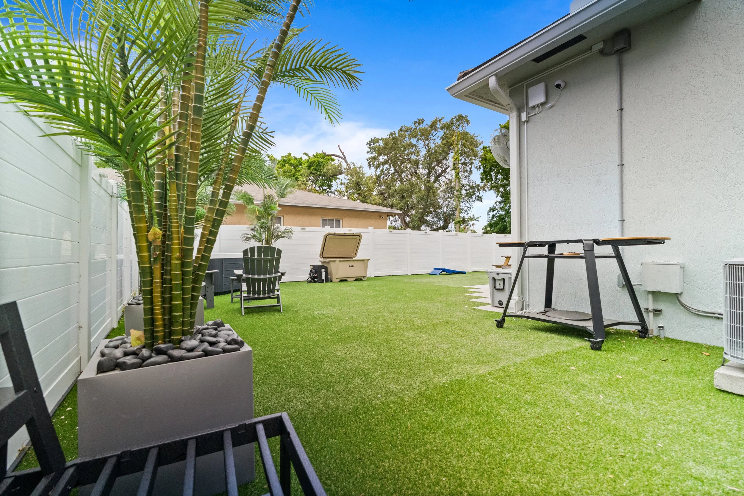 A backyard with artificial grass, potted palm trees, patio chairs, a storage box, and a white fence under a blue sky with some clouds.