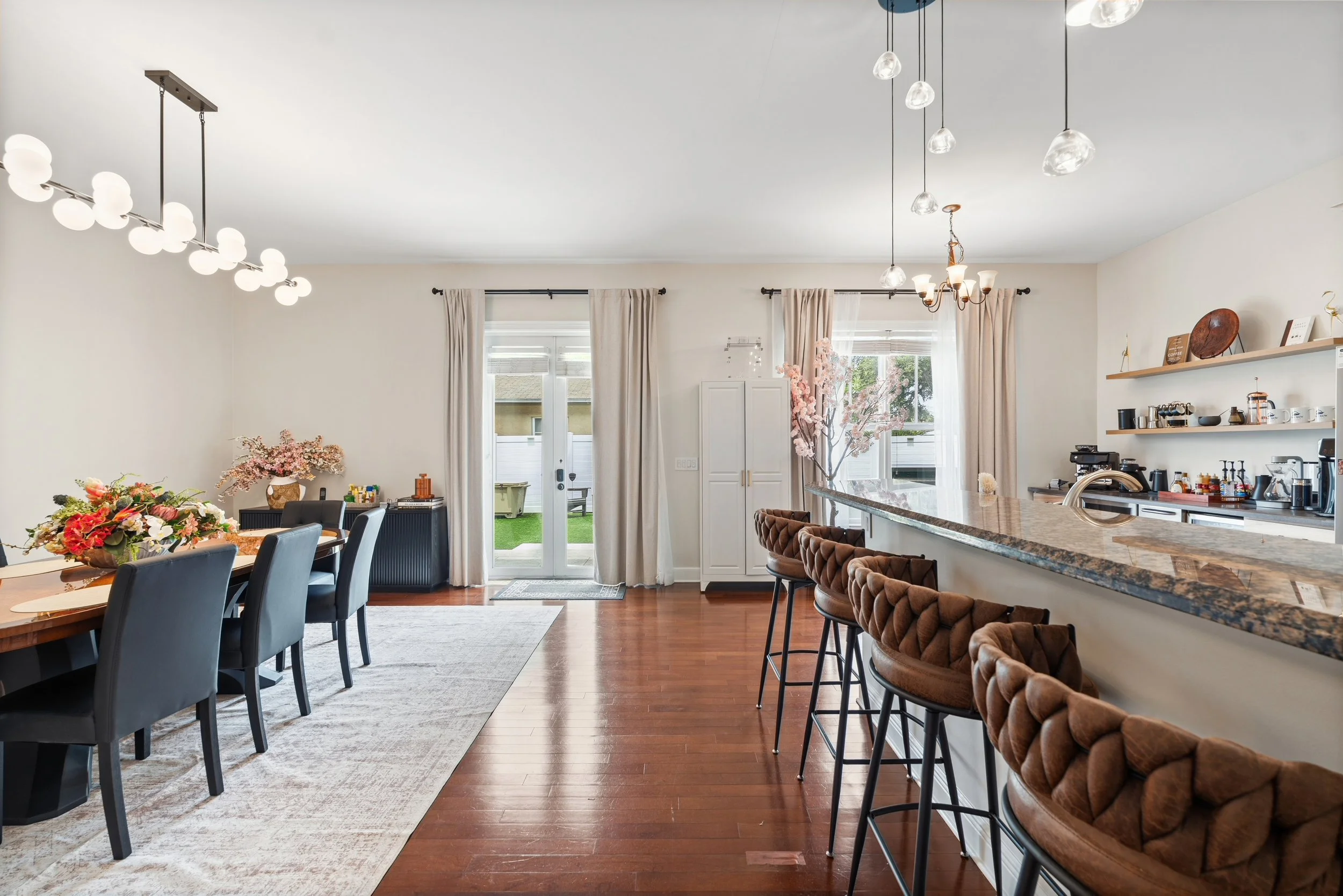 A modern living room featuring a dining table with black chairs, a floral centerpiece, and a kitchen bar area with brown tufted stools. The room has hardwood floors, large windows with beige curtains, and contemporary lighting fixtures, including a c