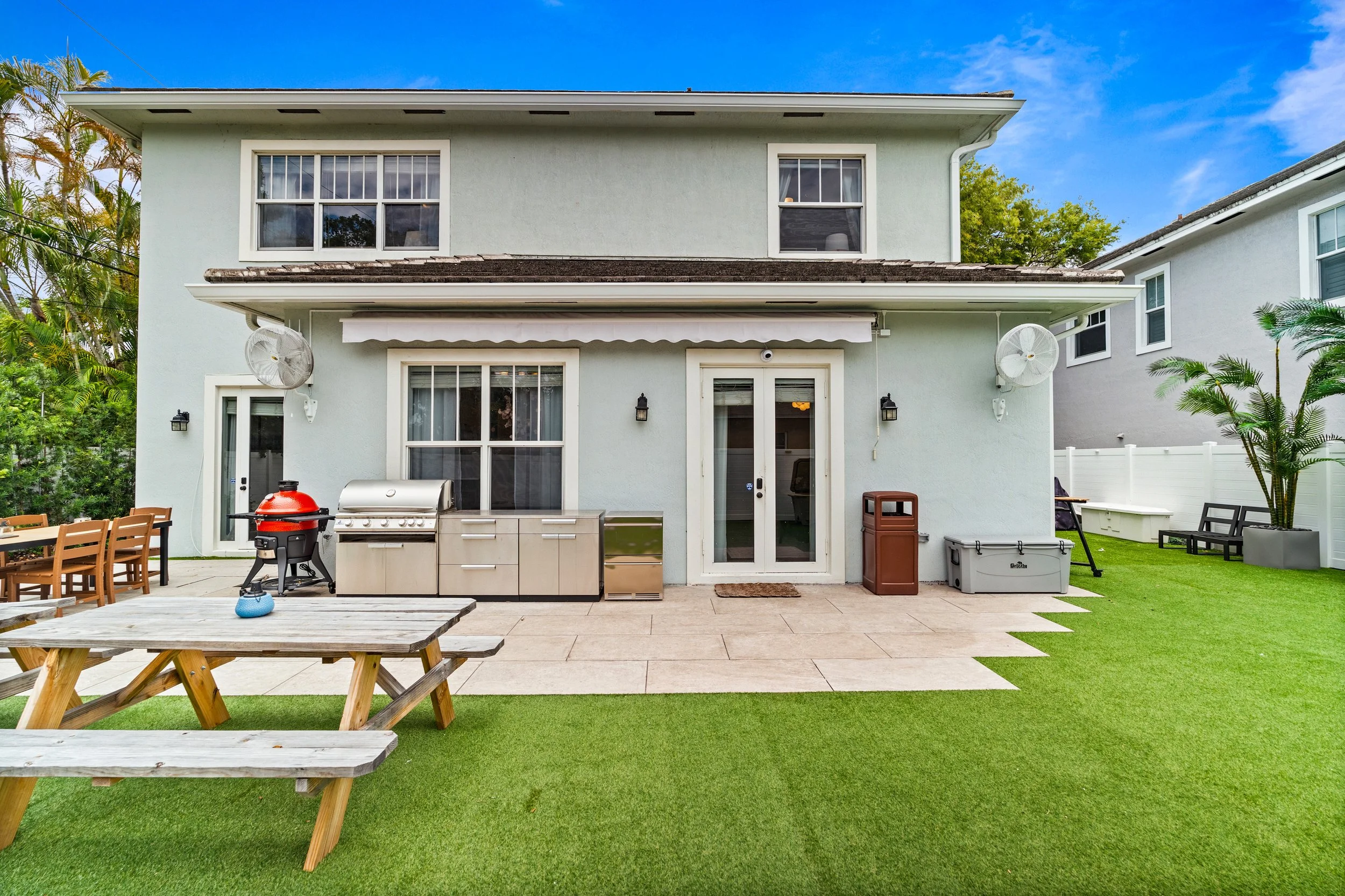 Backyard patio area with outdoor grill, wooden picnic table, and chairs. There are sliding glass doors, wall-mounted fans, and outdoor lighting fixtures on the house. The yard has grass and potted plants, with a white fence and neighboring house visi