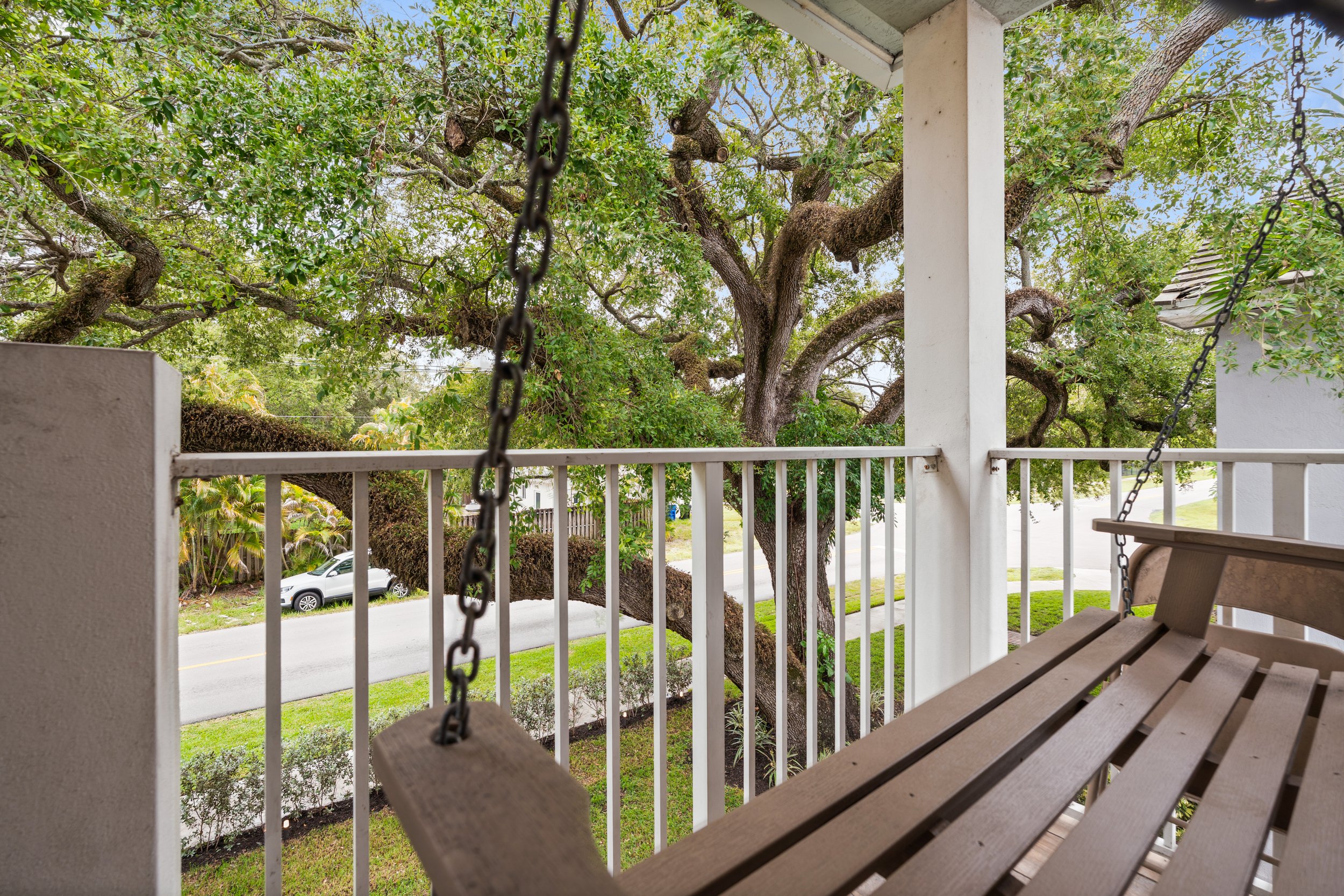 View from a porch showing a wooden swing in the foreground, a large tree with thick branches and green leaves, a white fence, and a street with a silver car parked beside it.
