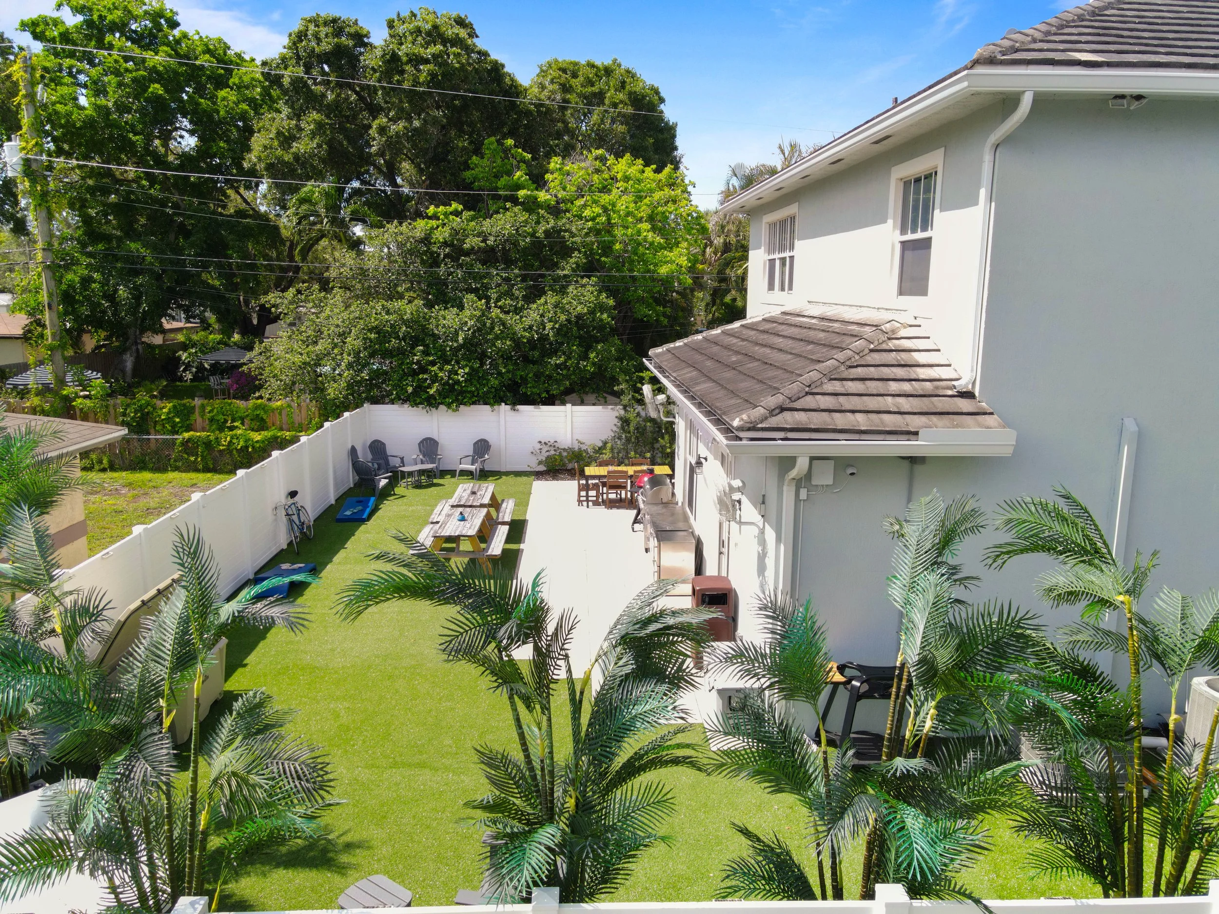 A backyard with a green lawn, patio furniture, and lush trees, enclosed by a white fence under a clear blue sky.