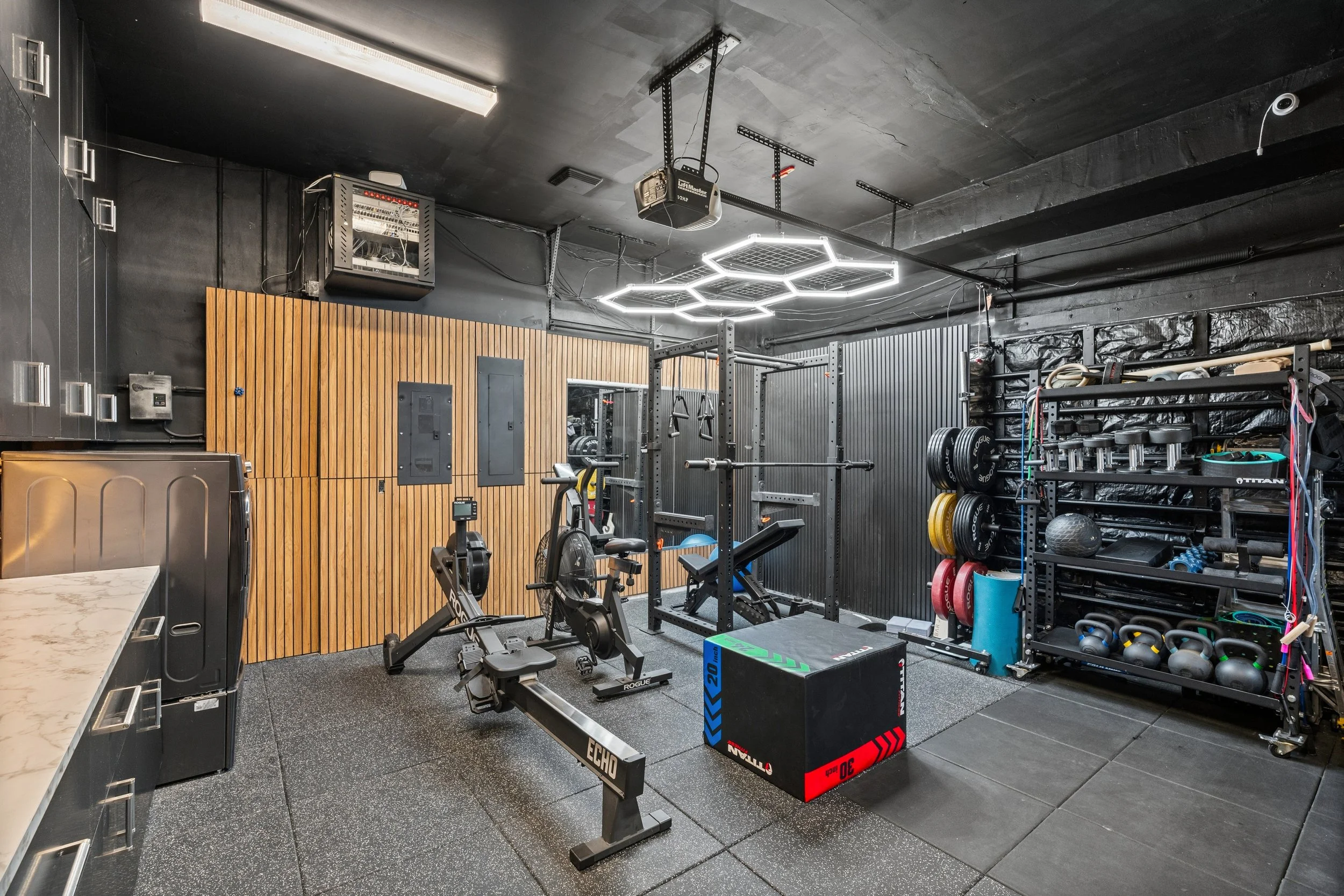 Modern gym with black walls and ceiling, wood paneling on one wall, and various workout equipment including squat racks, weight plates, kettlebells, exercise bikes, a punching bag, and a digital timer. Overhead lighting and hexagonal LED ceiling ligh