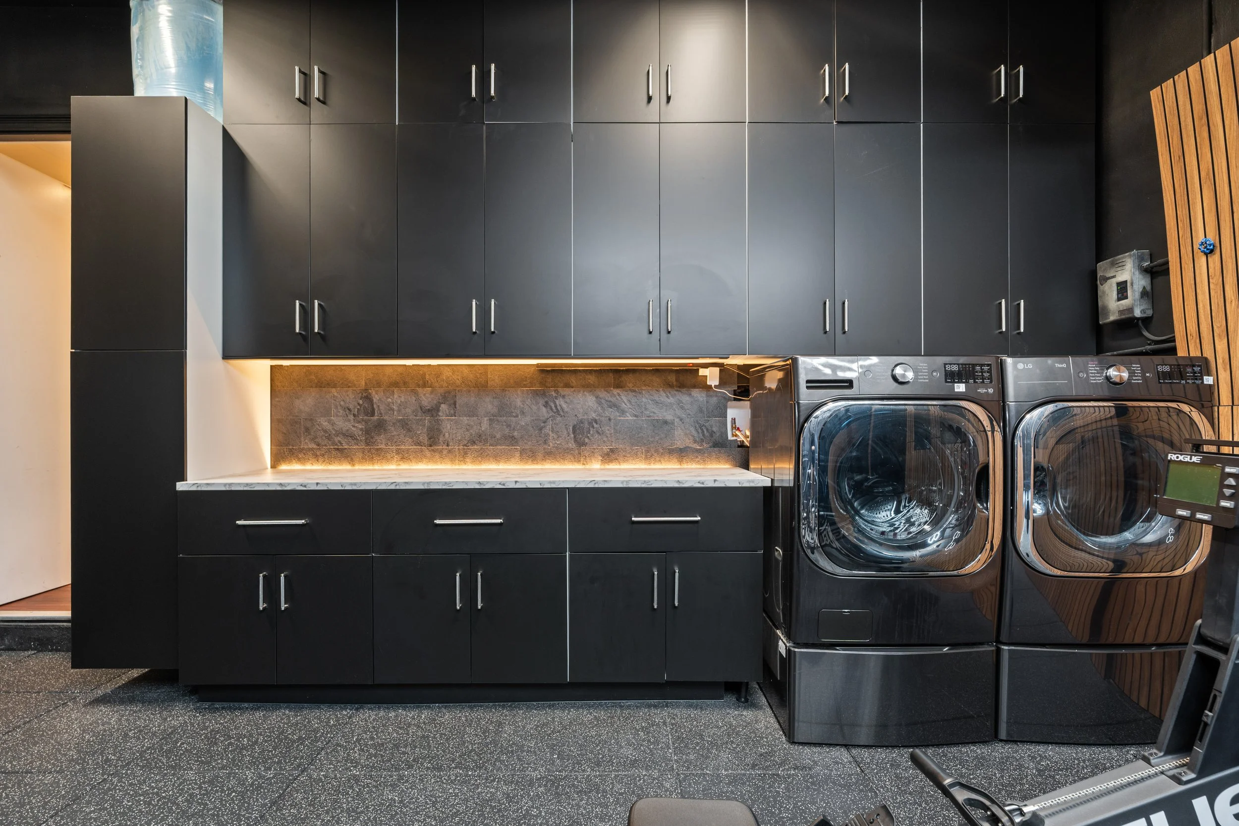 A modern laundry room with black cabinets, a marble countertop, and a tiled gray backsplash. Two front-loading washing machines are aligned under the cabinets, and a water heater is mounted on the wall to the right. There is speaker and some equipmen