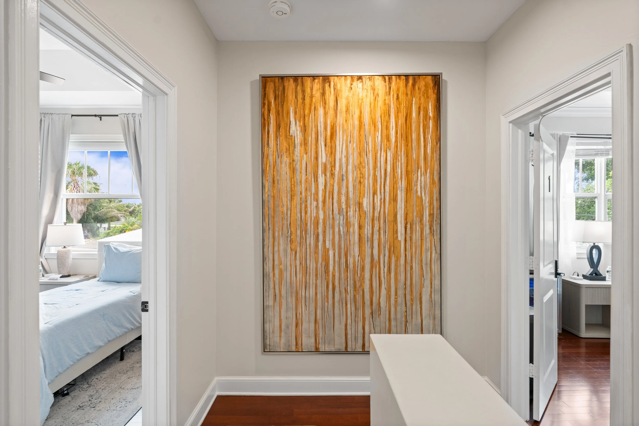 A hallway with a large abstract painting with orange, white, and brown vertical streaks on the wall. To the left, a bedroom with a bed, nightstand, and window with white curtains. To the right, part of another room with a window and a small dresser. 