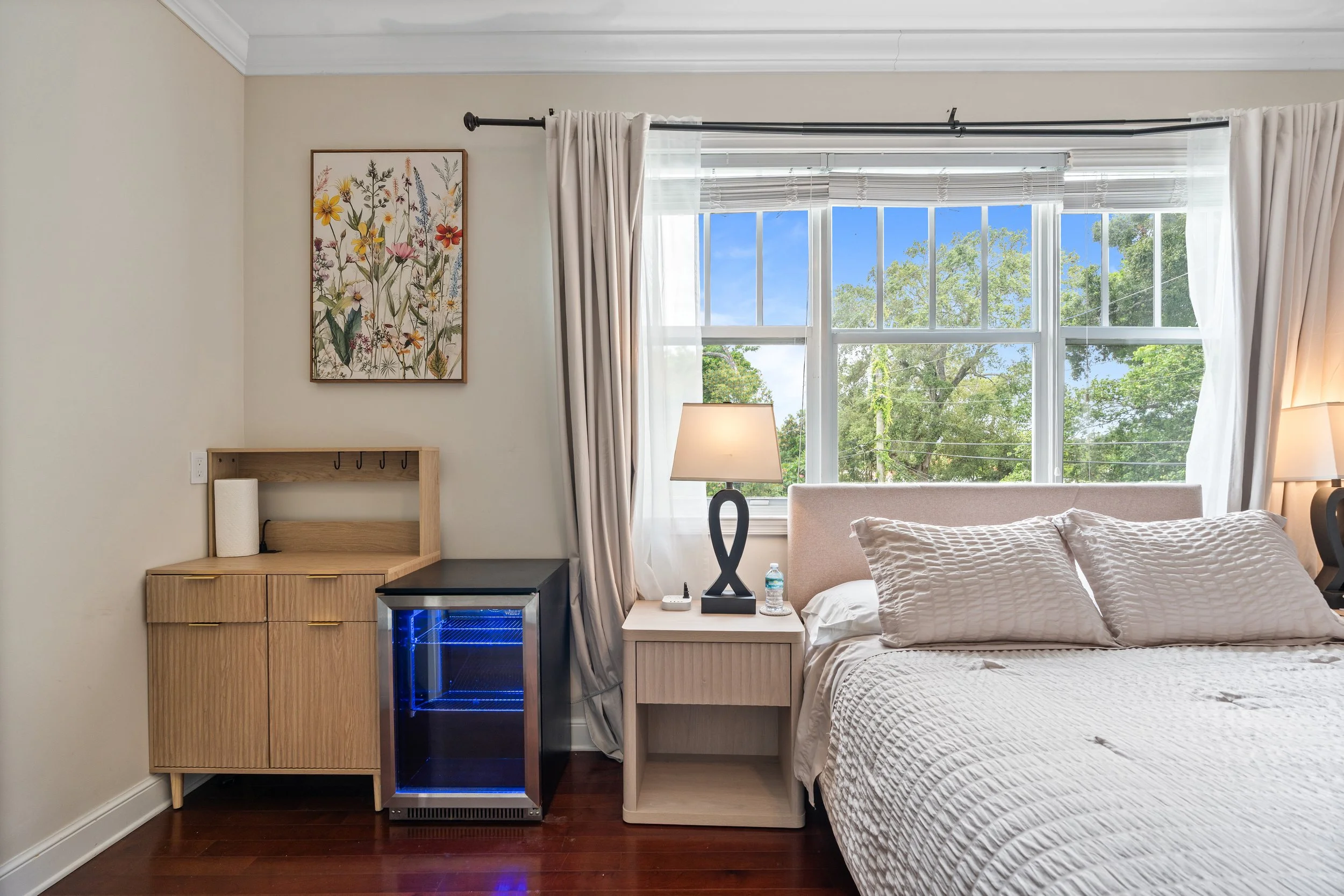 A bedroom with a large window showing green trees and a blue sky. There is a bed with a white quilt and pillows, a nightstand with a lamp, a small fridge, and a framed floral artwork on the wall.