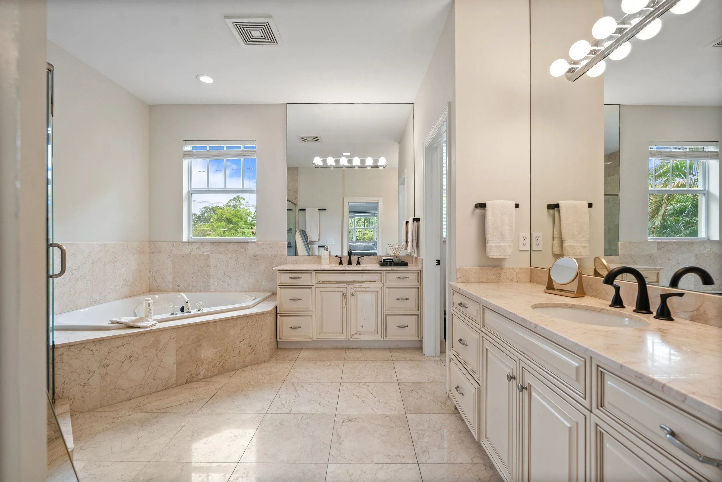 A spacious bathroom with a cream-colored marble tub and countertops, white cabinetry, a large mirror, black fixtures, illuminated vanity lights, two windows with blinds, and tropical greenery visible outside.