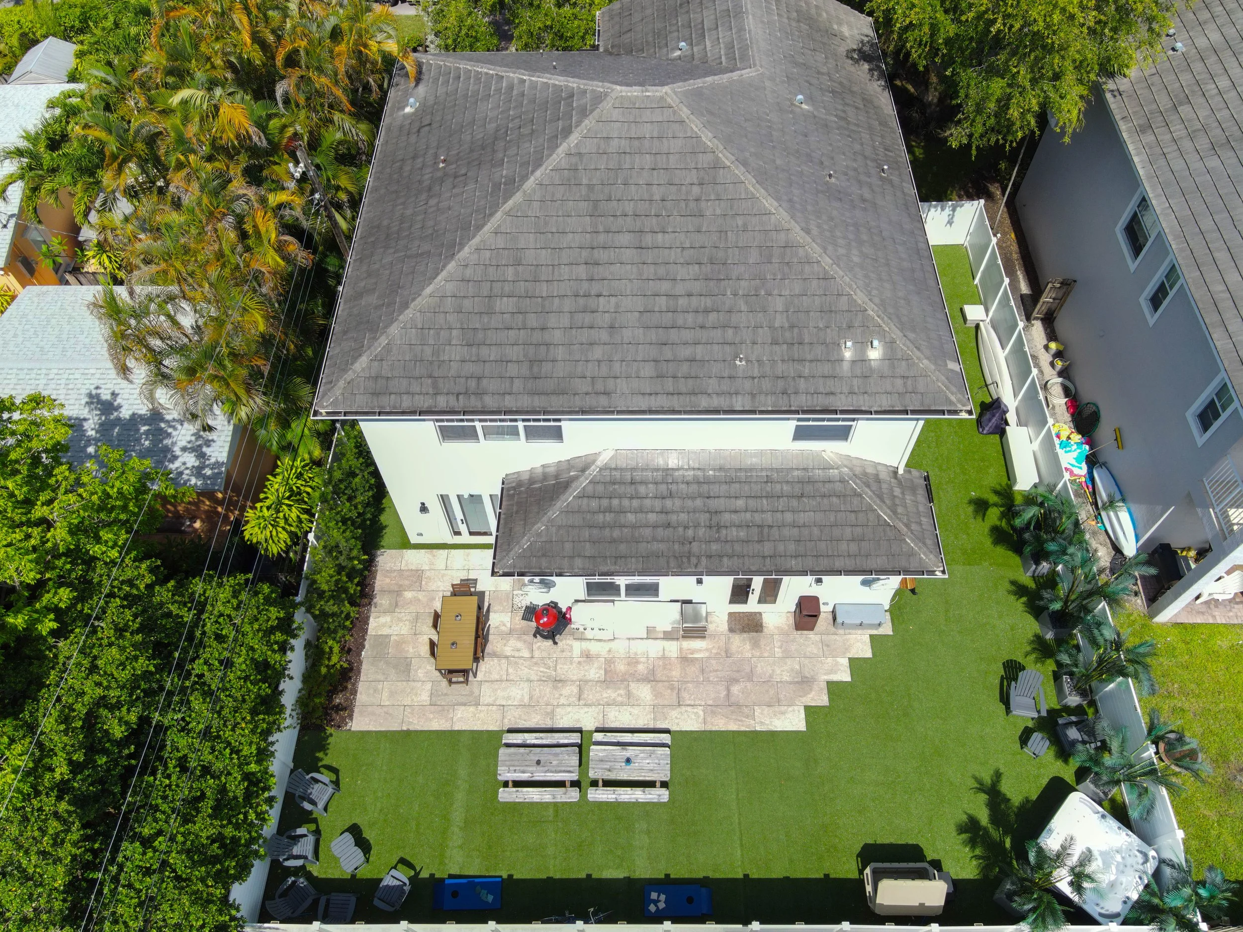 An aerial view of a backyard featuring a large house with a grey shingle roof, a concrete patio area with outdoor furniture and a barbecue grill, surrounded by a grassy lawn, various trees, and neighboring houses.