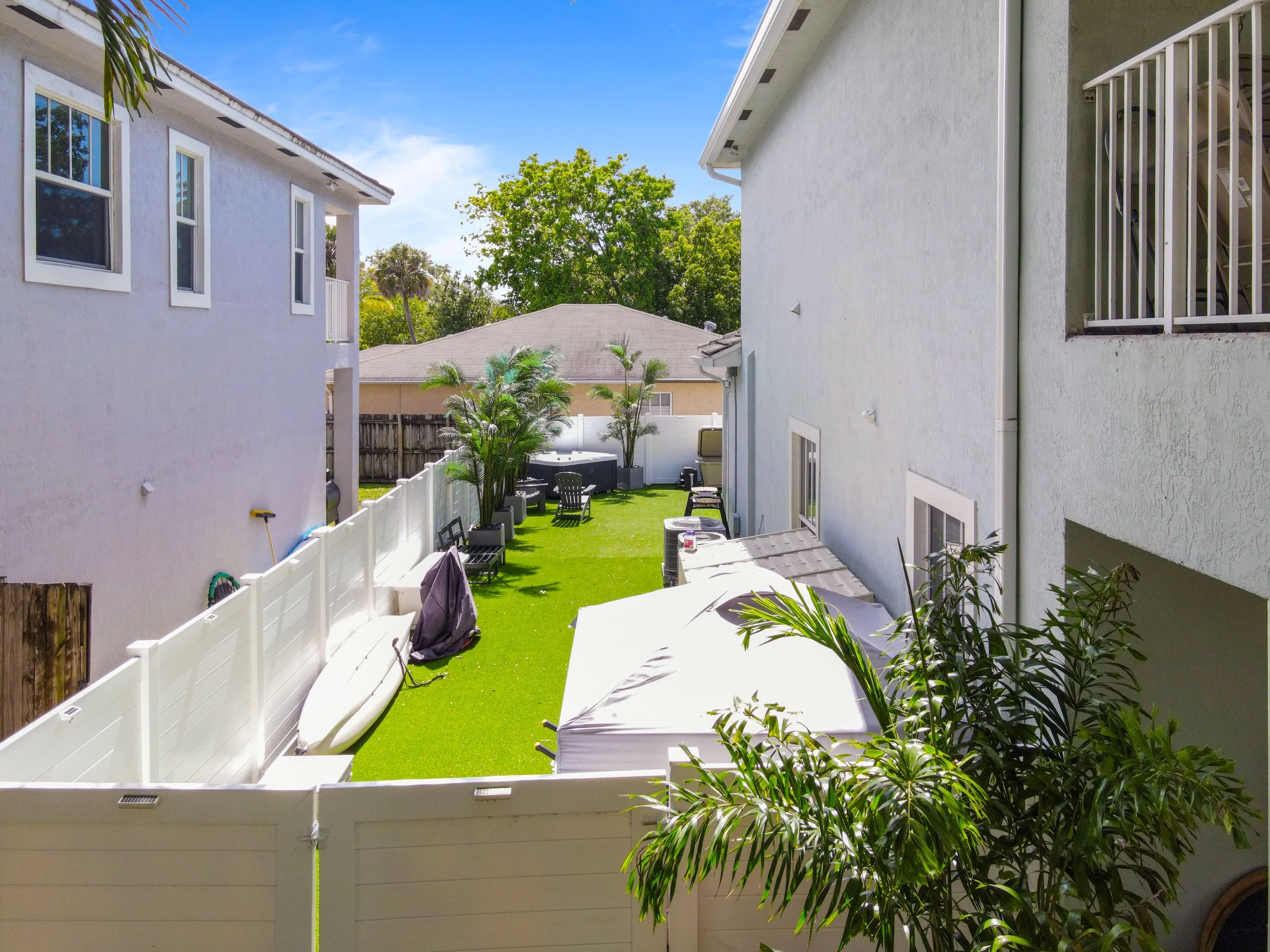 Backyard view with white fences, small palm trees, outdoor chairs, a shed, neighboring houses, and a clear blue sky.