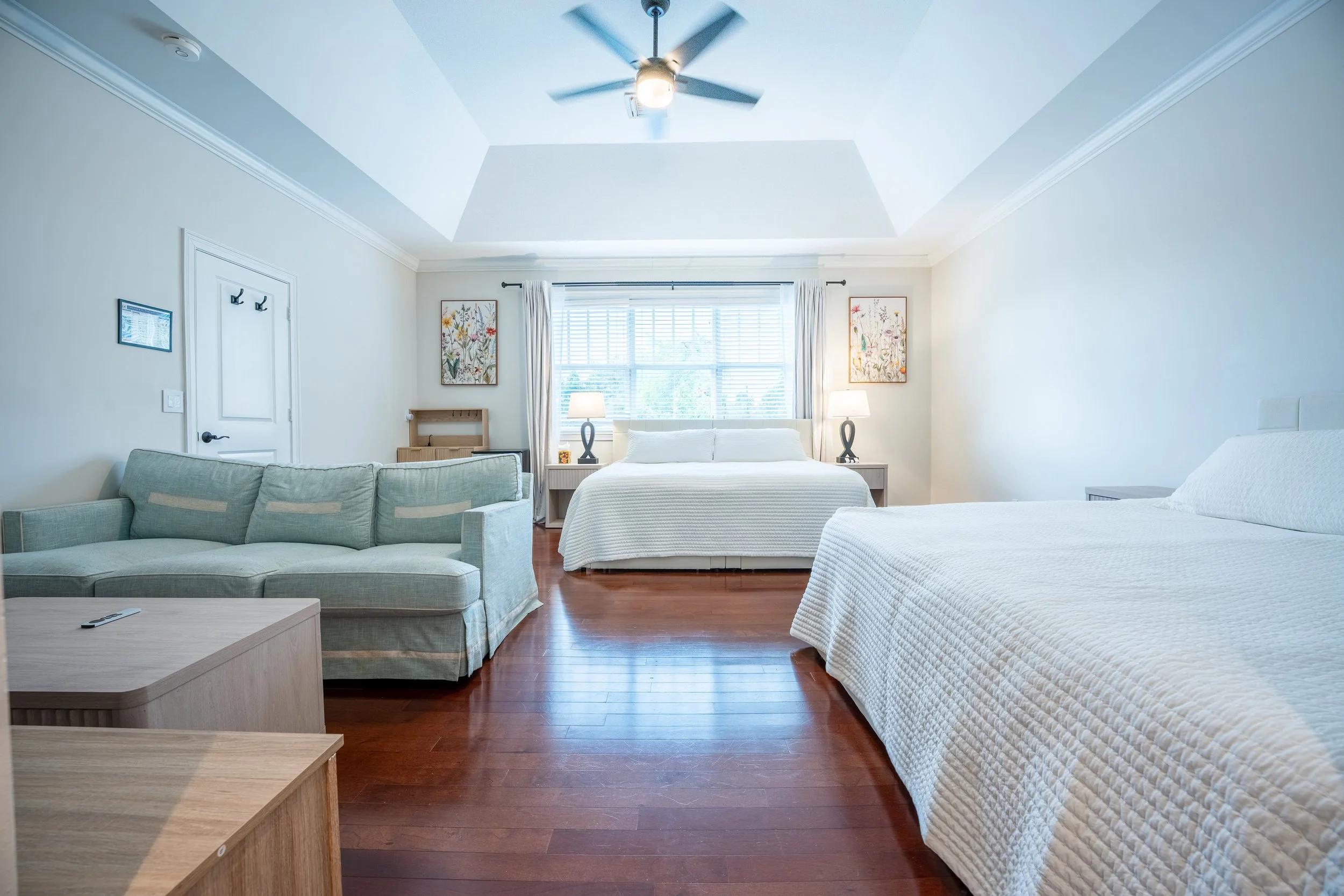 Bright hotel room with two beds, a sofa, a ceiling fan, large window with curtains, framed artwork, nightstands with lamps, and wooden flooring.