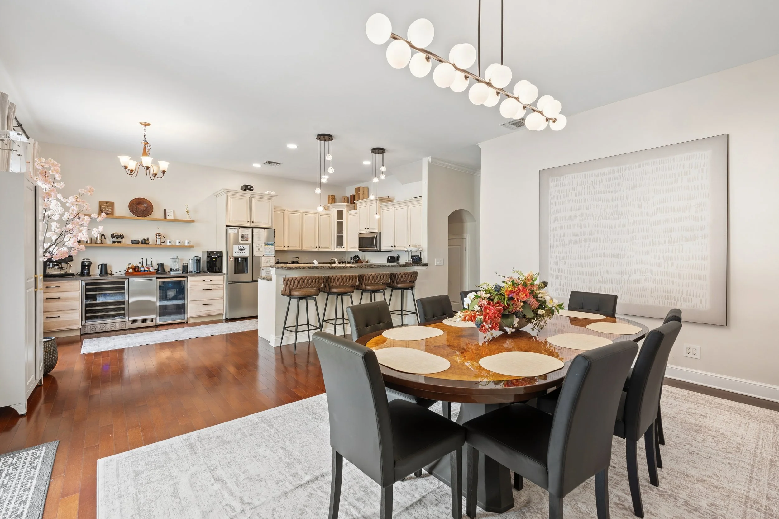 Interior of a modern open-plan kitchen and dining area with a round wooden table, black chairs, floral centerpiece, kitchen appliances, white cabinetry, and decorative lighting fixtures.