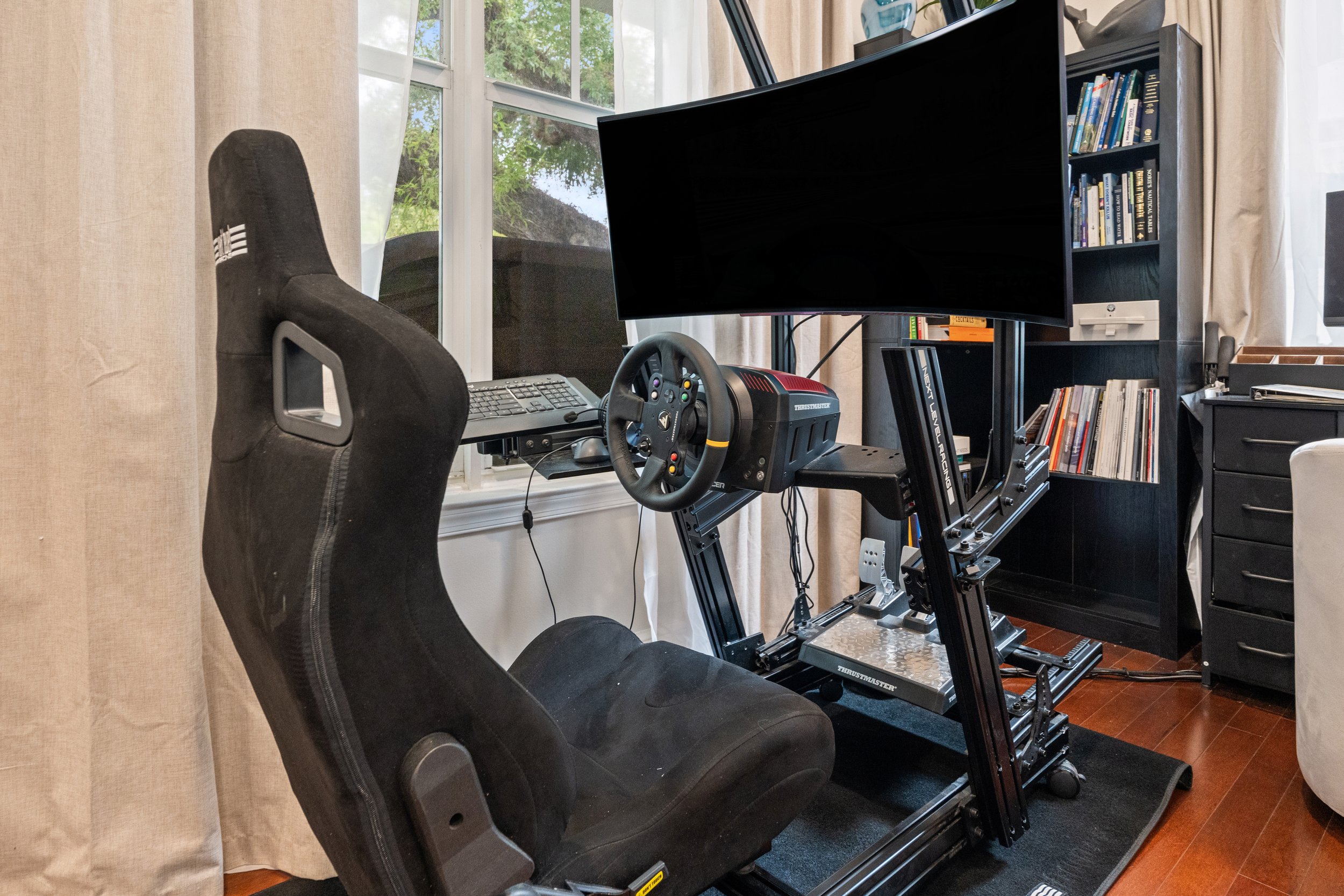 A home office with racing simulator setup including a large curved monitor, racing steering wheel, and a gaming chair. There is a bookshelf with books and a window with curtains and greenery outside.