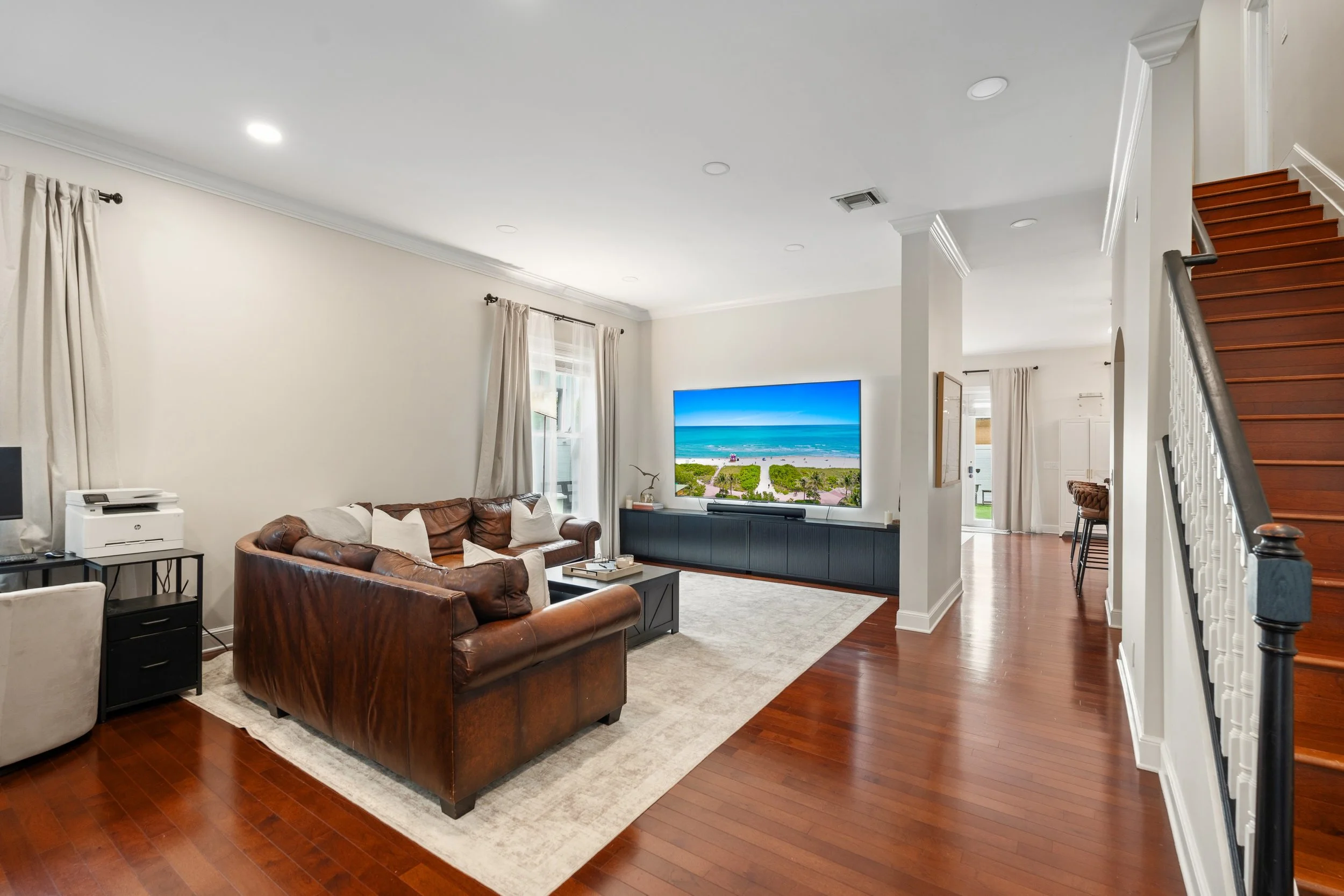 Living room with leather sofas, TV showing beach scene, windows with curtains, staircase, hardwood floors, and modern decor.