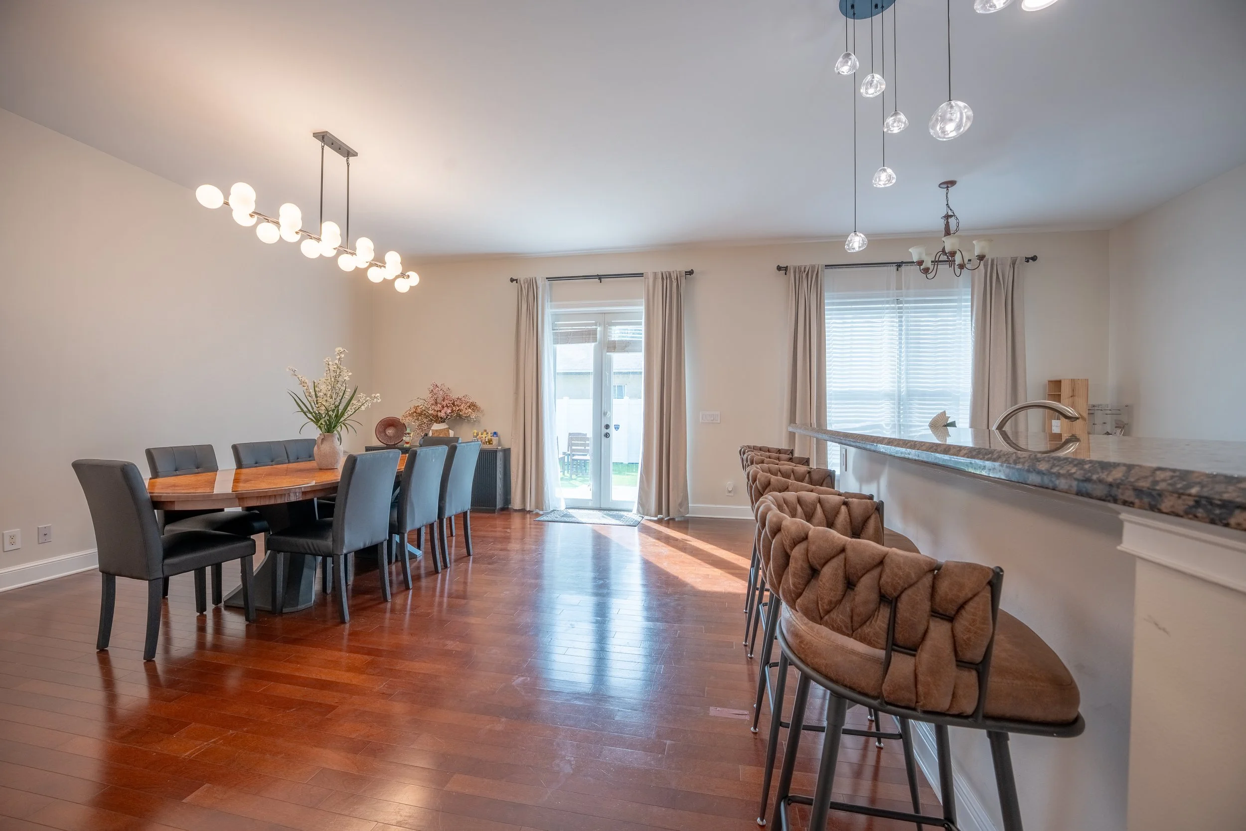 Dining area with a wooden table, eight black chairs, floral decorations in vases, large windows with curtains, and sliding doors leading to an outdoor space; kitchen bar with beige cushioned stools and pendant lights overhead.