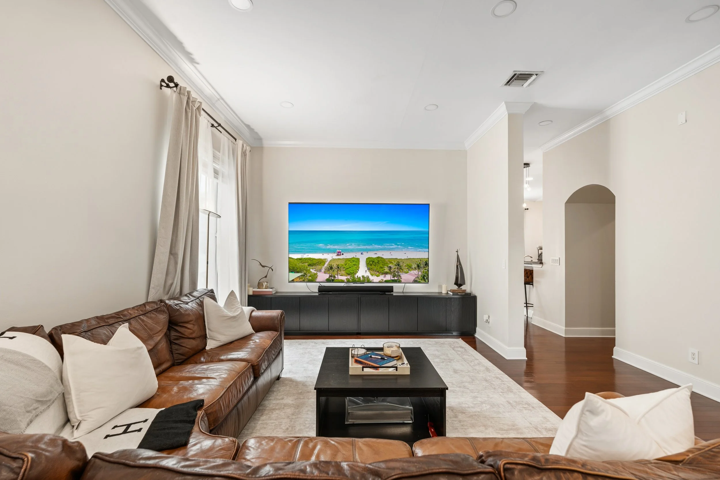 Living room with a tan leather sofa, white pillows, a dark wood coffee table, large window with curtains, and a flat-screen TV showing a beach scene.