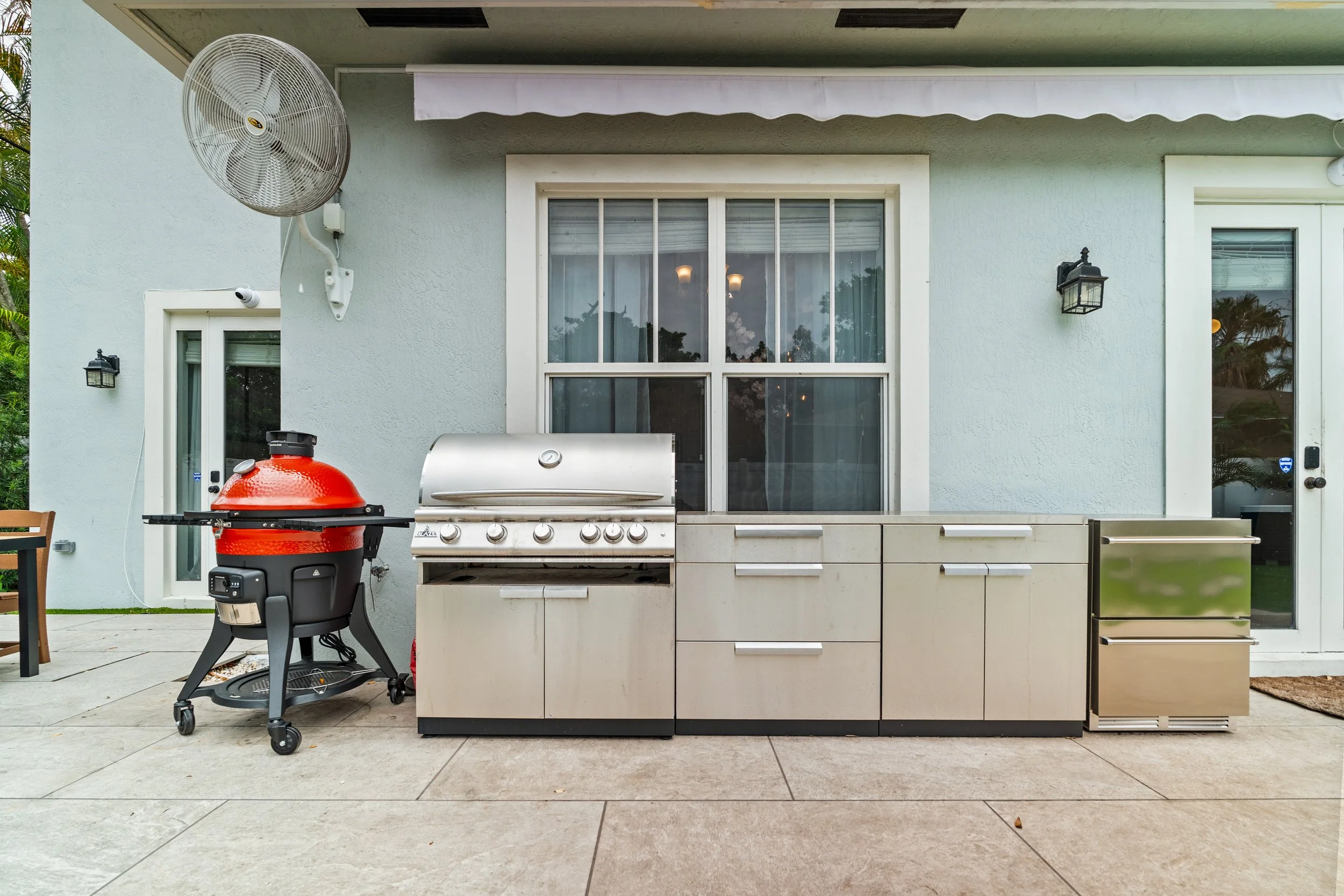 Backyard patio with an outdoor grill and kitchen area in front of a house with light blue walls, large sliding glass doors, and exterior lighting fixtures.