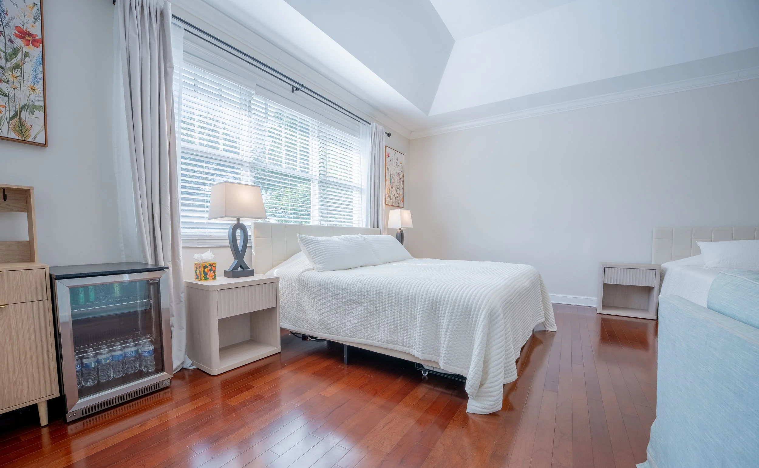 A bright bedroom with white walls, two beds with white bedding, a window with blinds, and wooden floors. There are two matching lamps on nightstands and framed floral artwork on the wall.