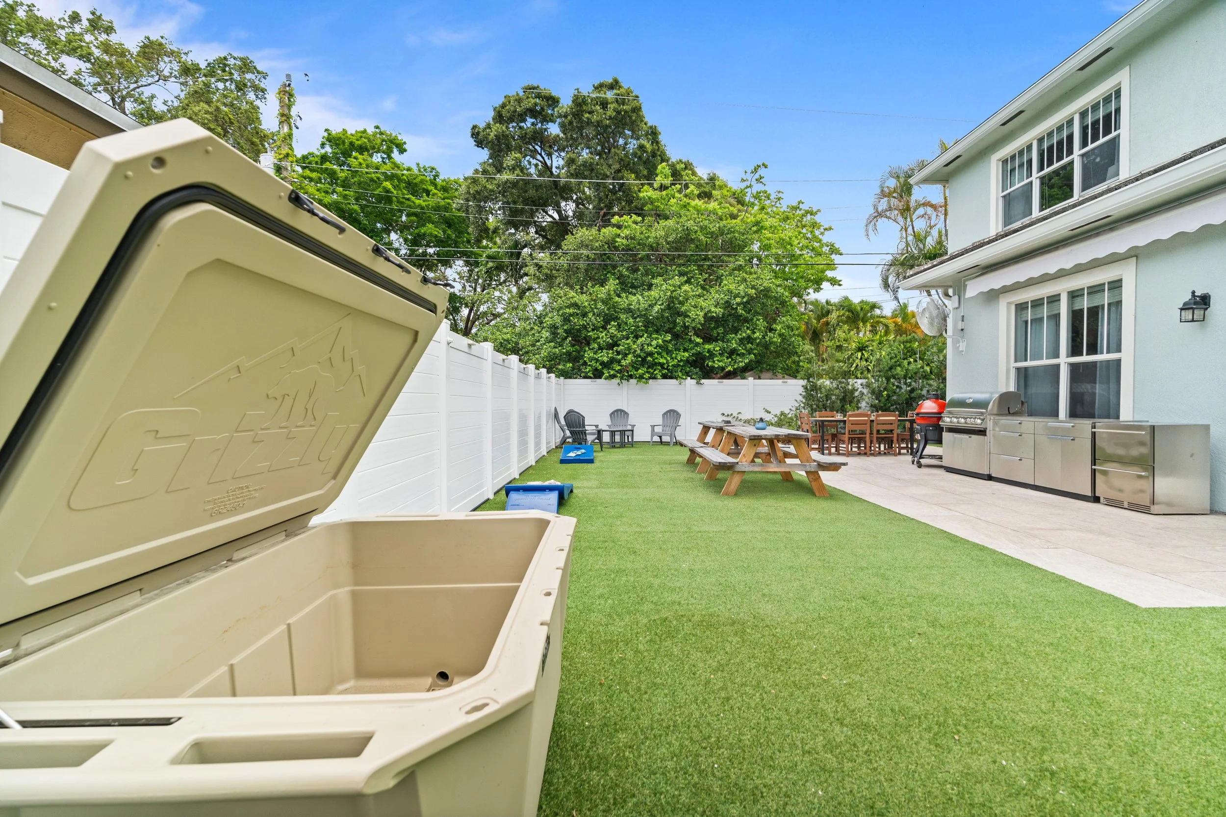 Backyard with artificial grass, white fence, picnic table, Adirondack chairs, grill, and bright blue sky.