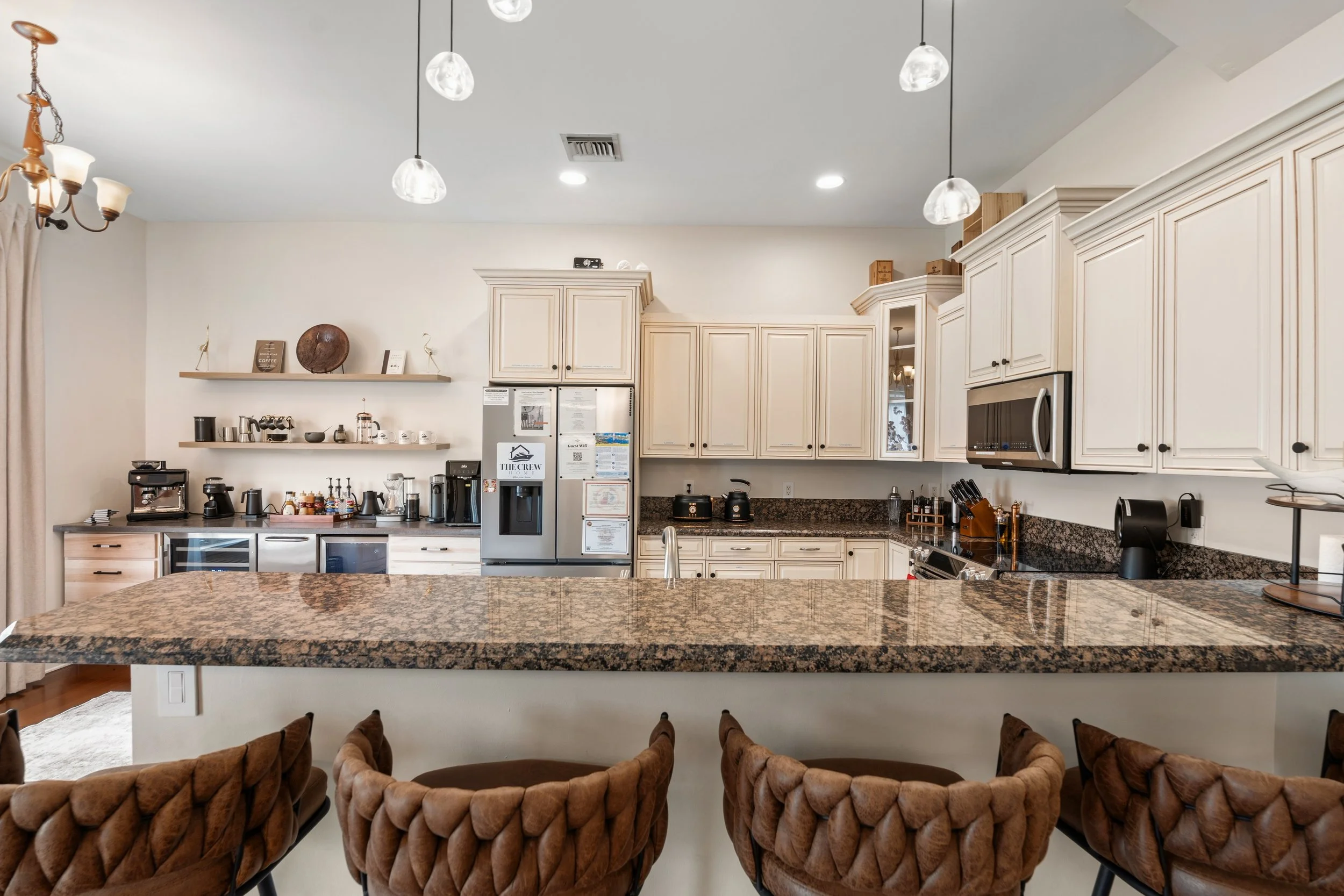 Modern kitchen with cream-colored cabinets, granite countertops, coffee equipment, and hanging pendant lights over the island. There are chairs in the foreground.