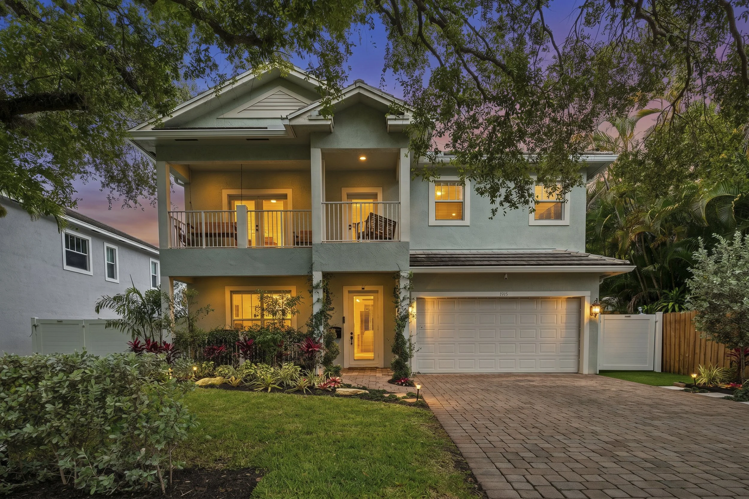Two-story house with front yard garden illuminated by outdoor lights at dusk, with a brick driveway leading to a garage and trees around the property.
