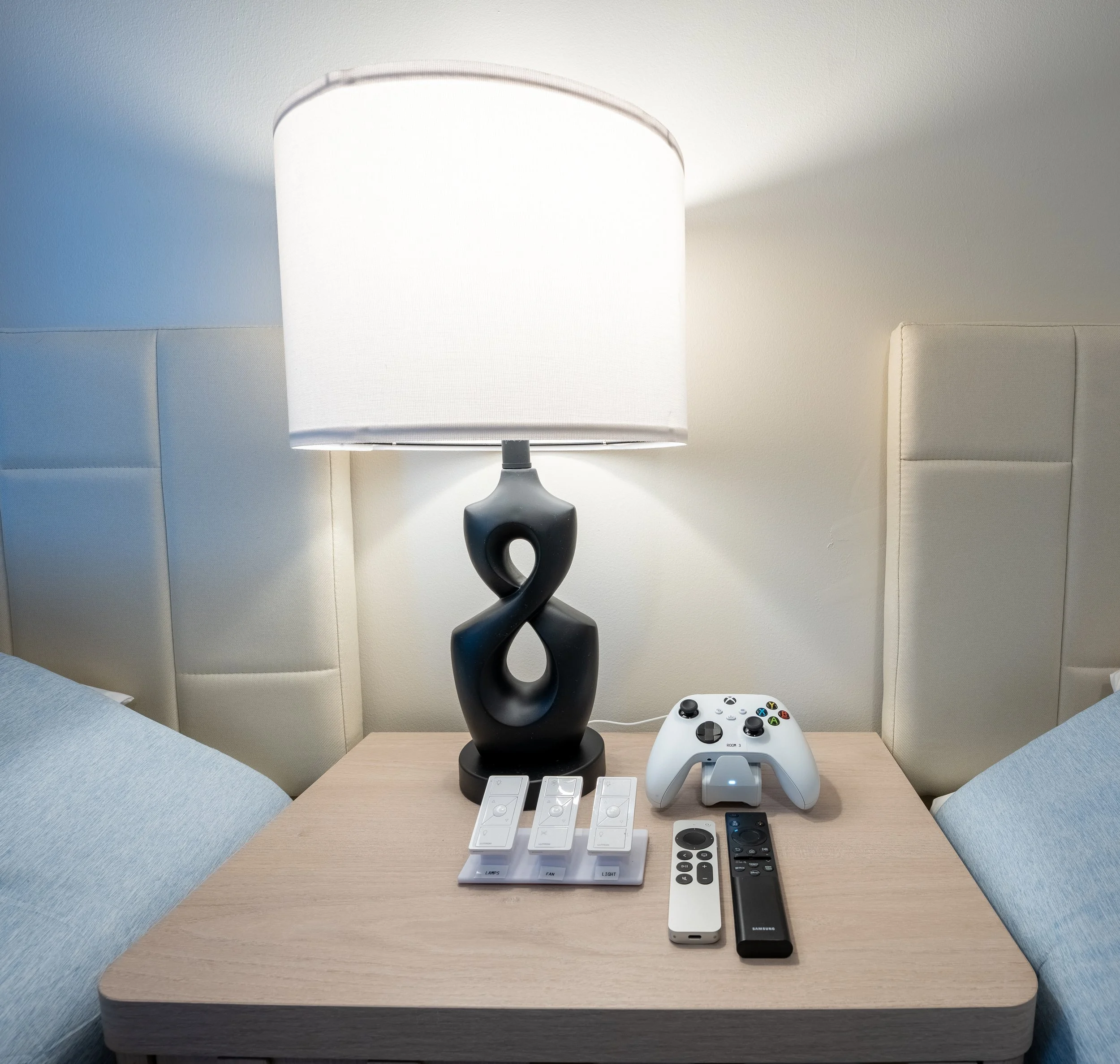 A bedside table with a black abstract lamp, a white Xbox controller, various remote controls, and smart light switches.