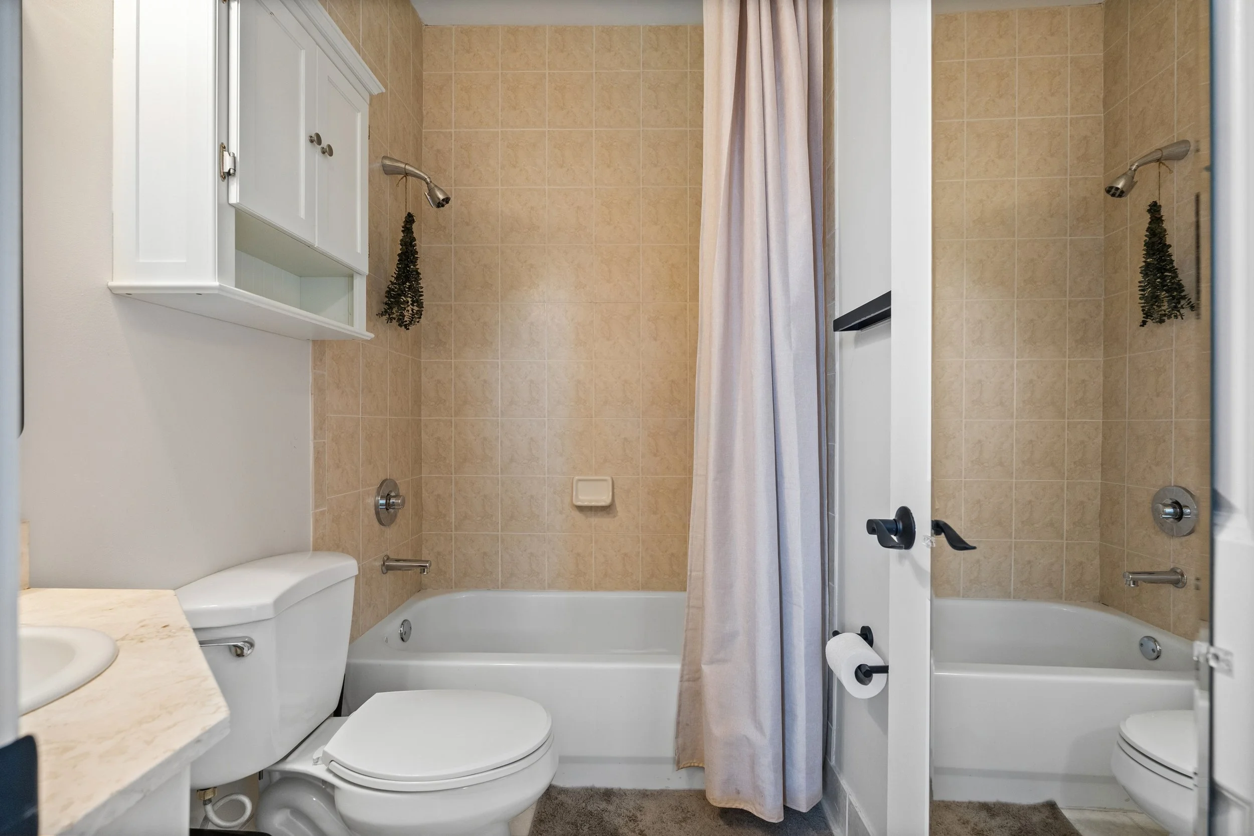 A small bathroom with beige tiled walls, a white toilet, and a shower with a beige curtain. There is a white cabinet mounted above the toilet, a small white soap dish on the shower wall, and a white towel on the left side. A sink countertop with a ma