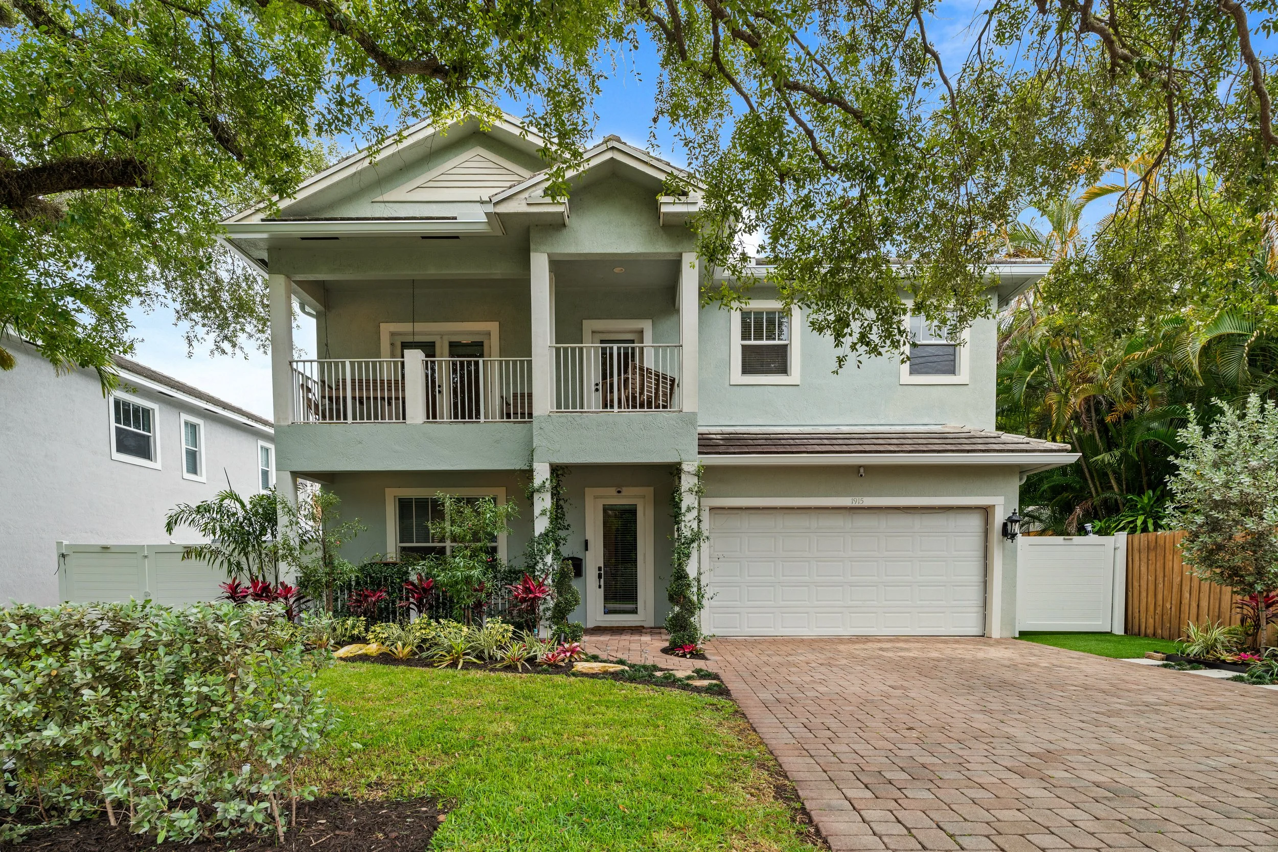 A light green, two-story house with a white garage door, front porch, and balcony. The house is surrounded by a garden with shrubs and colorful plants, and a brick pathway leads to the front door. Tall trees partially shade the house, and the sky is 