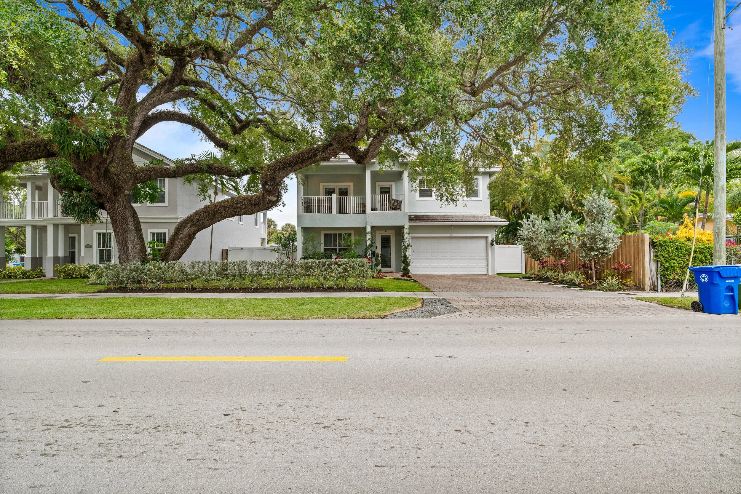 A residential street scene with a house partially obscured by a large tree with sprawling branches. The house is white with a balcony and a garage, surrounded by greenery. A blue sky with some clouds is visible, and there is a blue recycling bin on t