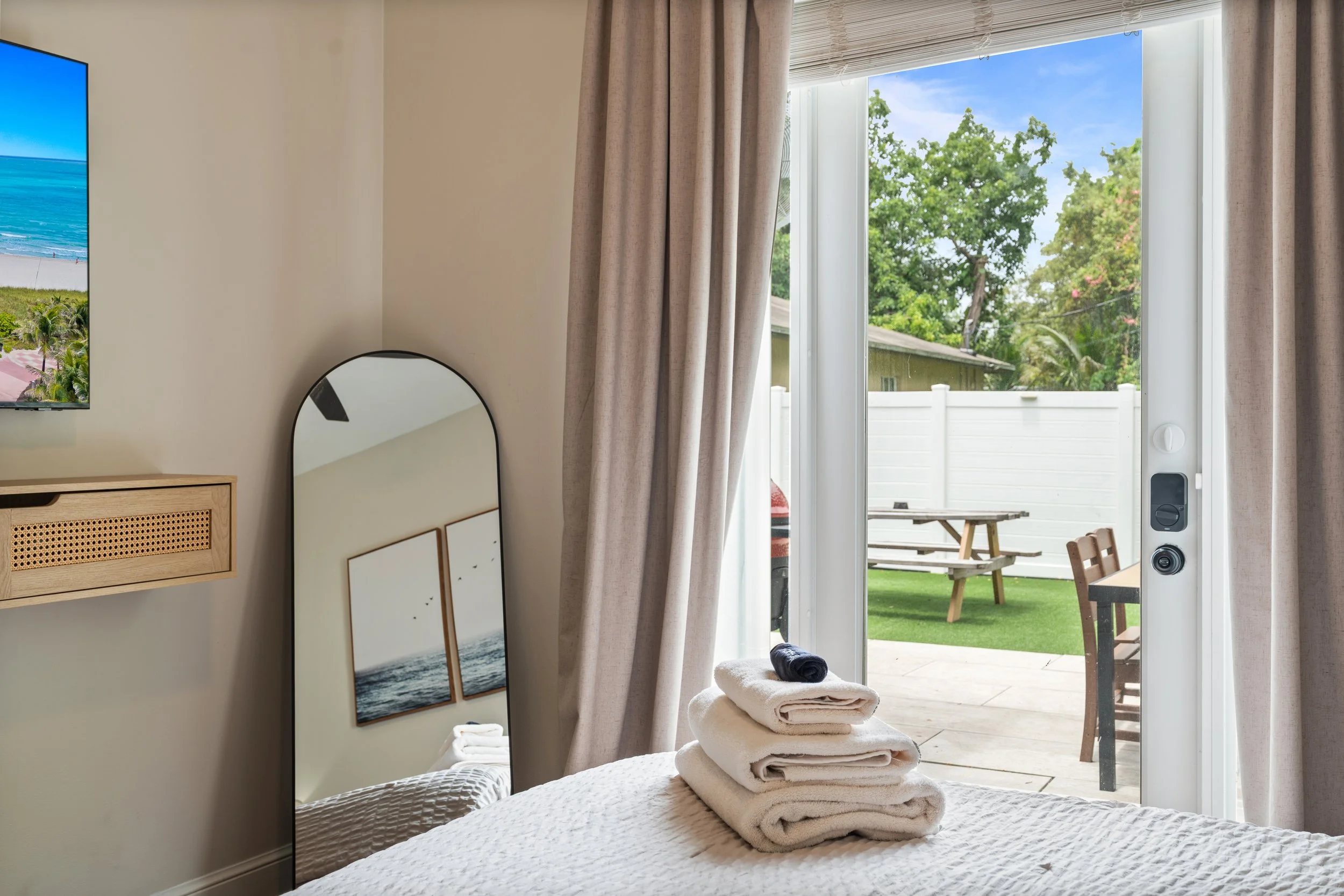 A bedroom with beige curtains open to a backyard patio with a picnic table, lush green trees, and a white fence, and a staircase leading outside.