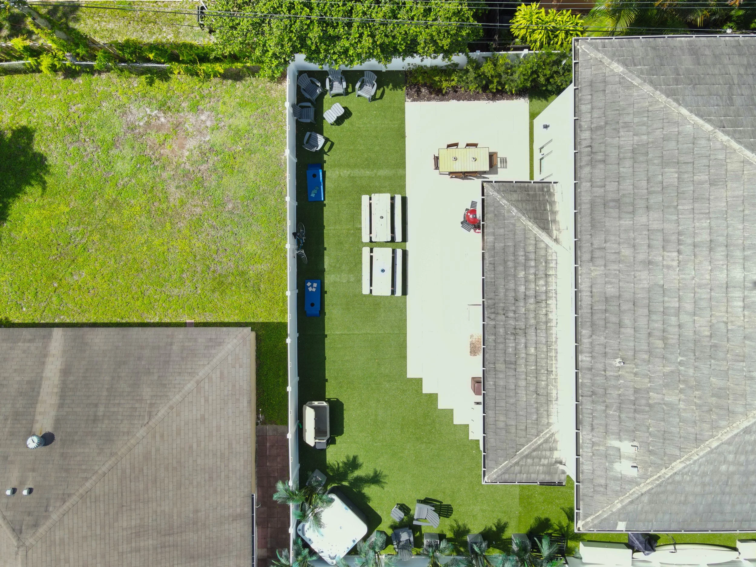 An aerial view of a backyard divided into a grassy area with chairs and a barbecue grill, and a paved patio with outdoor furniture, partially surrounded by a white fence and neighboring rooftops.