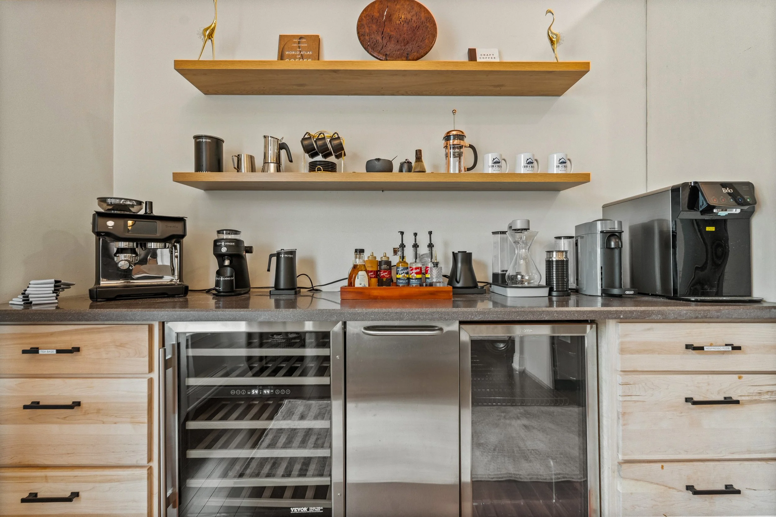 A kitchen countertop with coffee-making appliances, mugs, spice bottles, and two wooden shelves with decorative items and books.