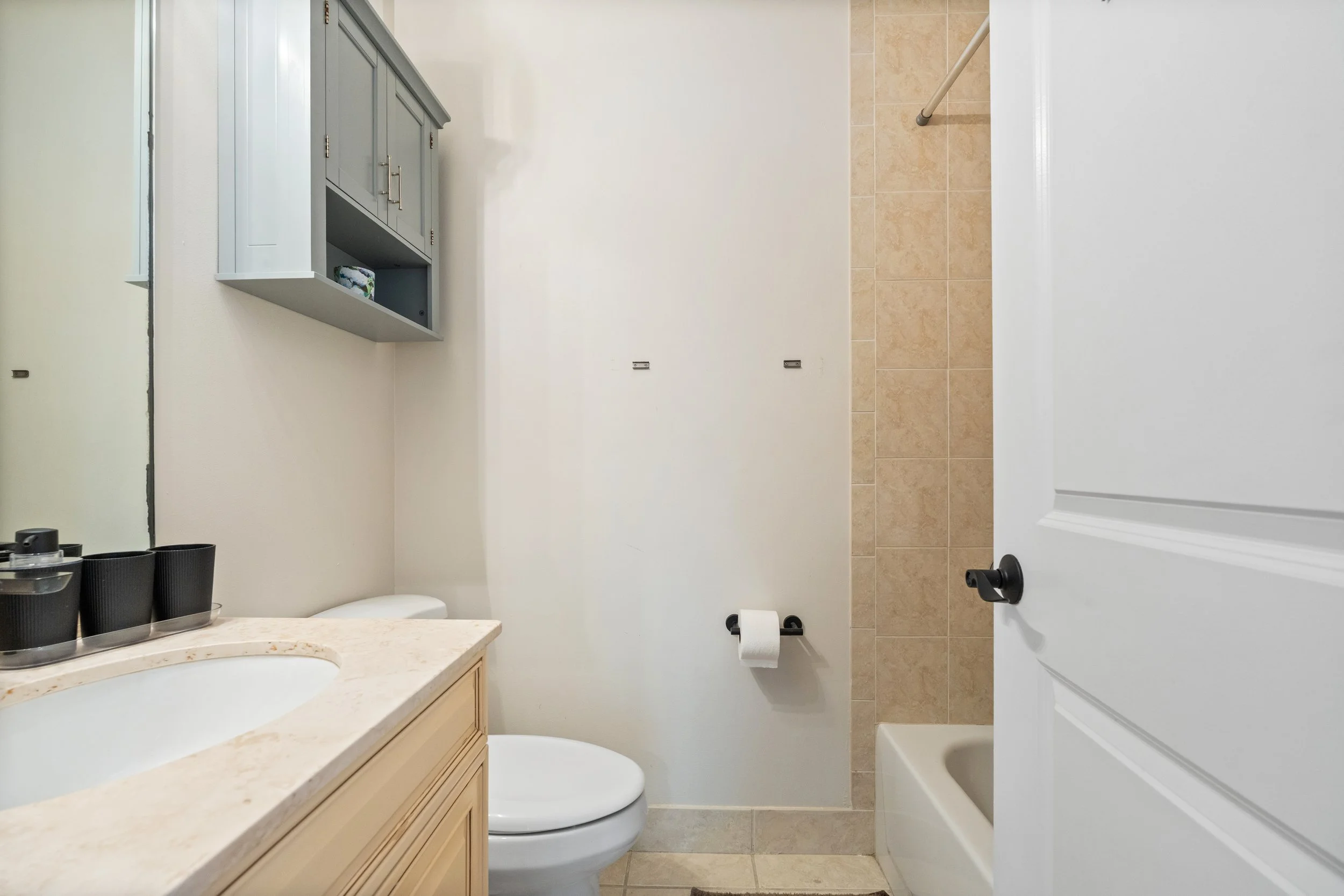 A small bathroom with beige tile flooring and a white wall. There is a white door on the right, a toilet with a toilet paper holder, black cups on a marble countertop, a wall-mounted cabinet, and a shower area.