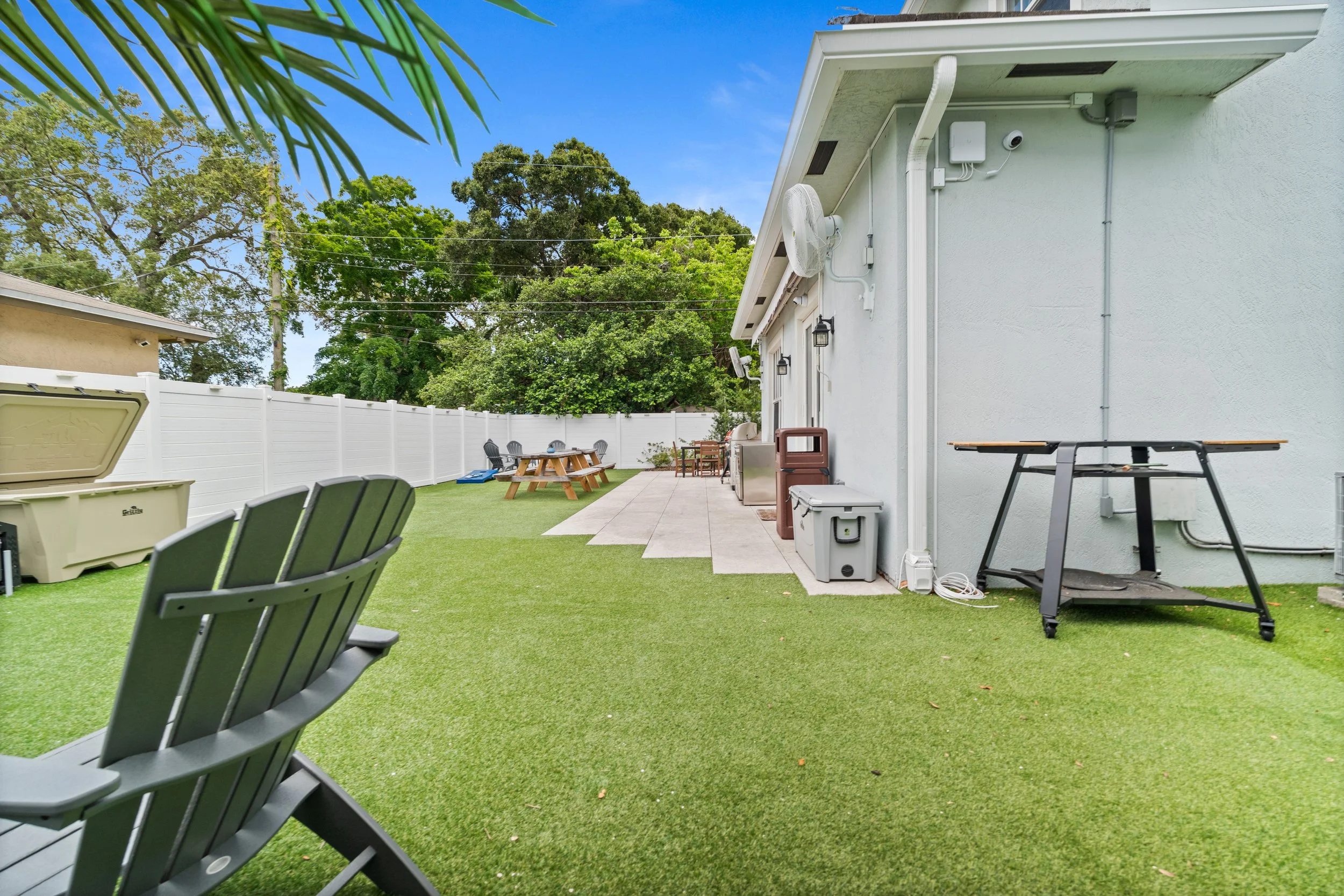 Backyard patio with artificial grass, outdoor seating, a picnic table, and various outdoor appliances and furniture, enclosed by a white fence under a blue sky with trees in the background.