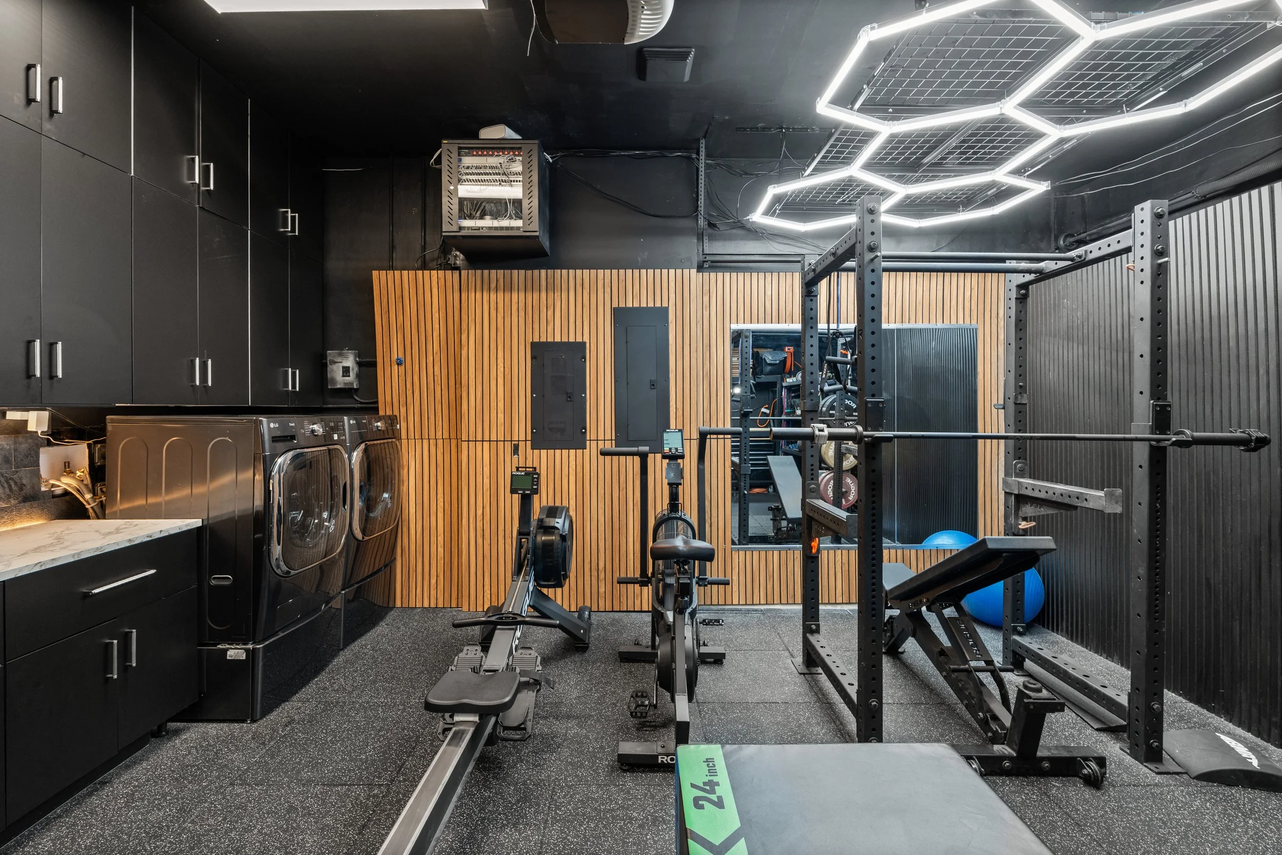 A black and gray home gym with fitness equipment, black cabinets, a wooden wall, LED ceiling lights, and a laundry area with washing machines.